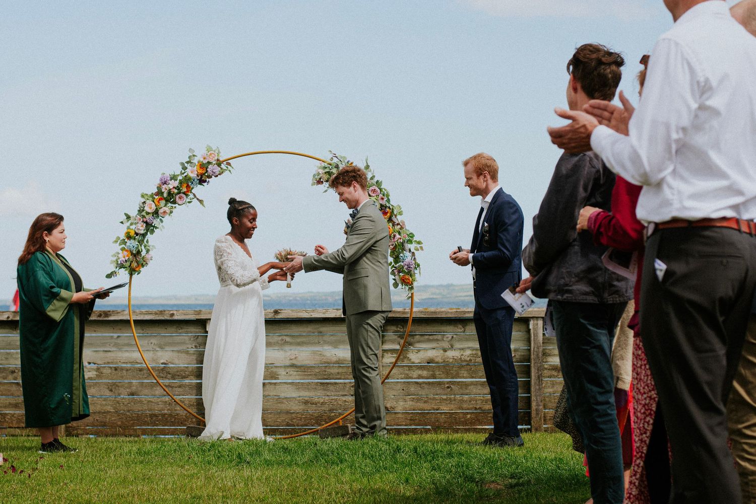 Wedding ceremony taking place outdoors under a floral arch with guests gathering on a grassy lawn.