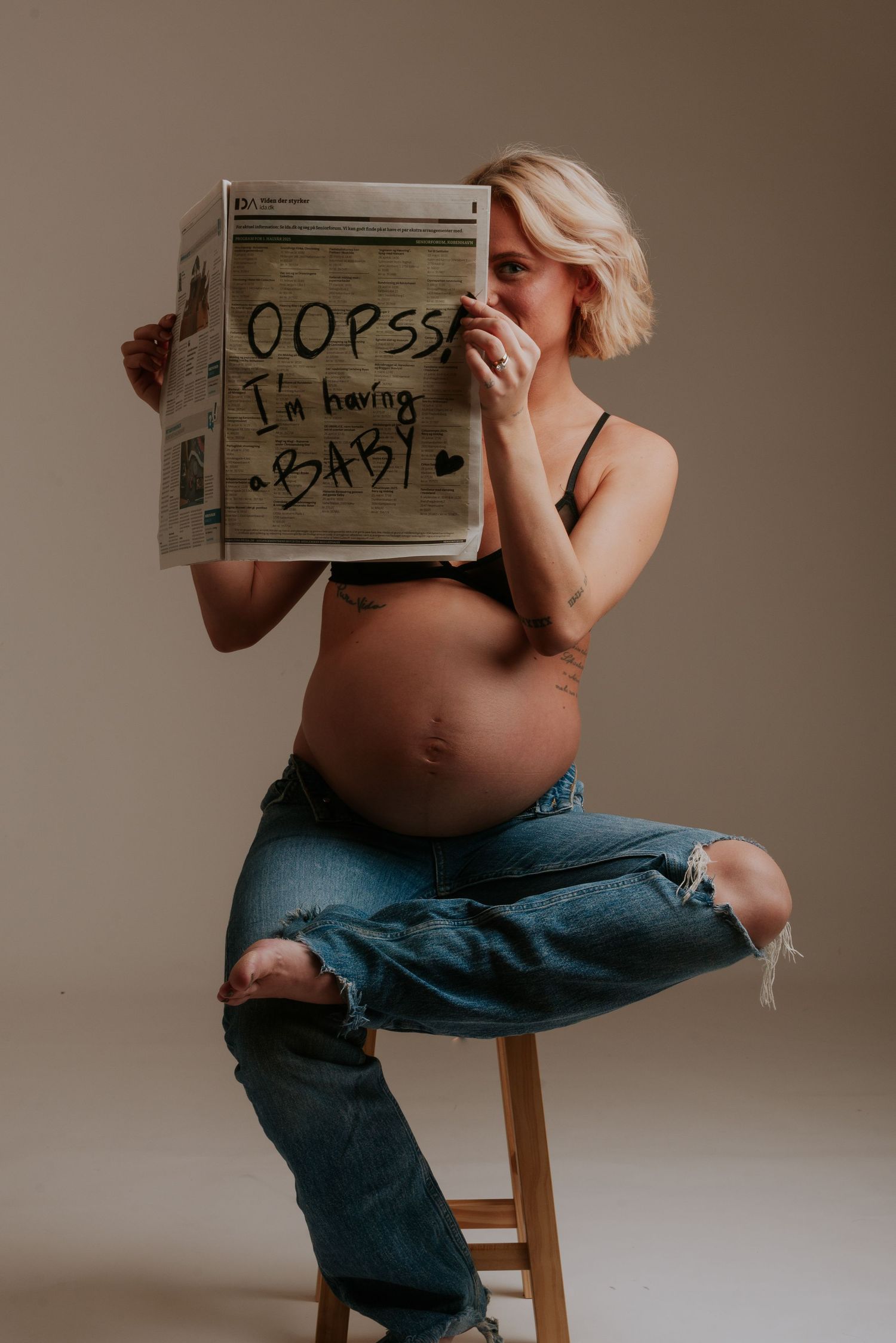 Creative maternity photo of an expectant mother in ripped jeans holding a sign saying 'oops baby' while sitting on wooden stool.