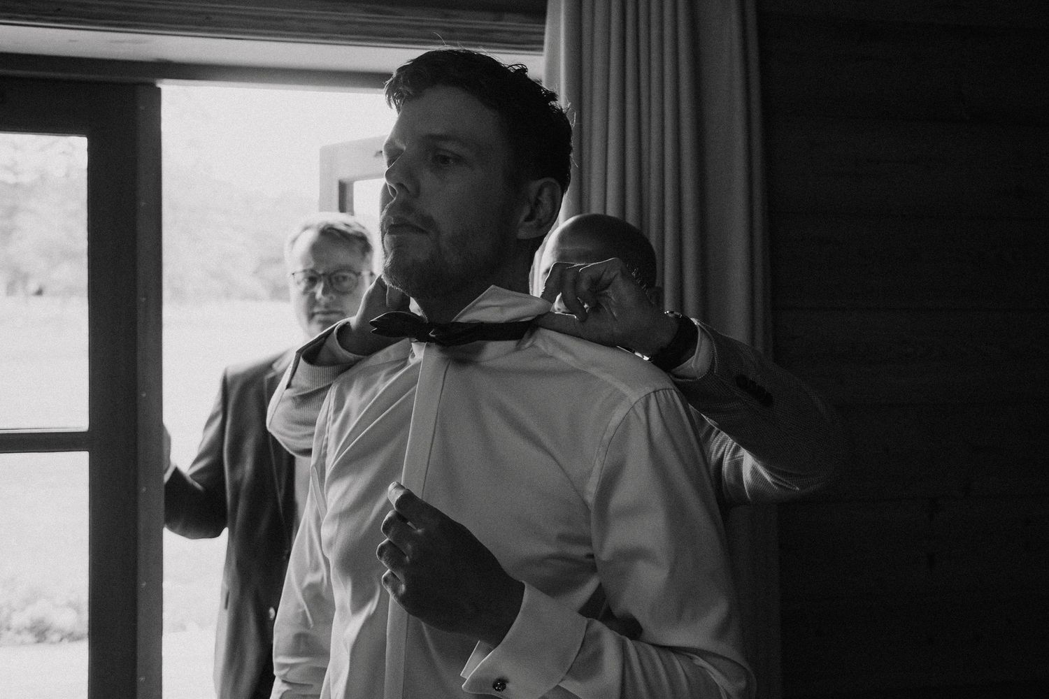 A groom getting ready in a white shirt as someone helps adjust his attire.