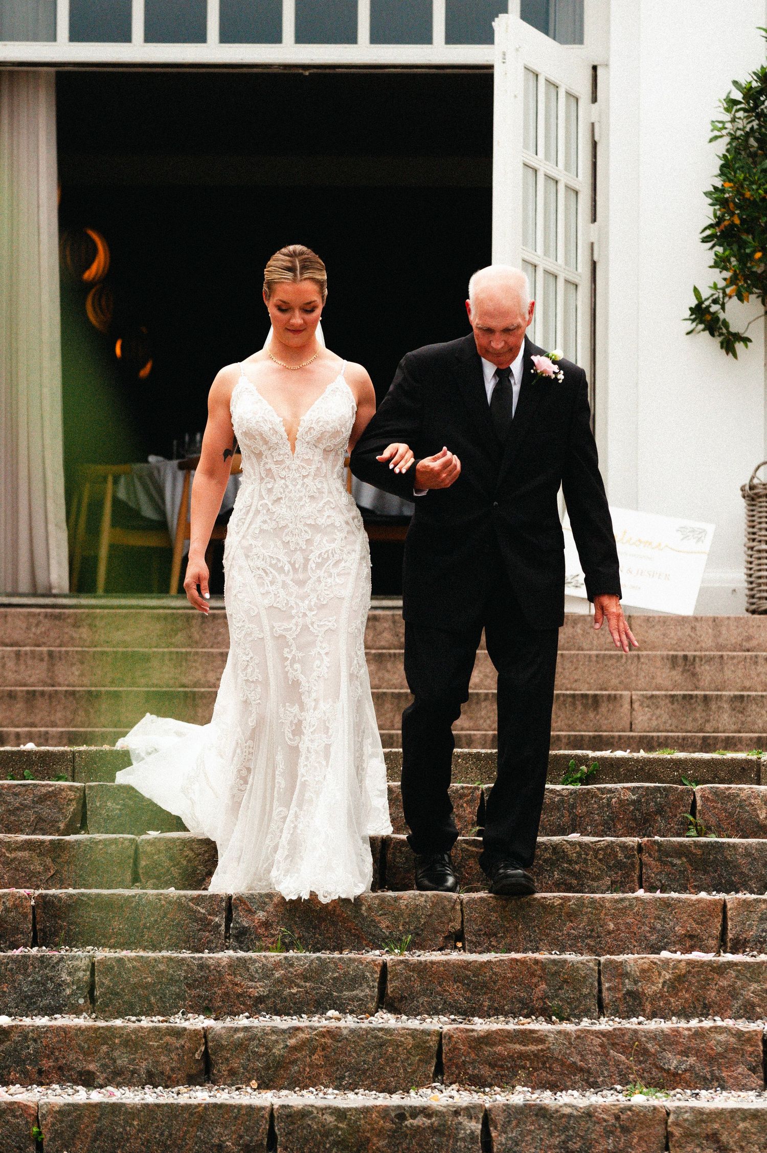 A bride in a white lace gown descends stone steps with an escort in a black suit during a wedding ceremony.