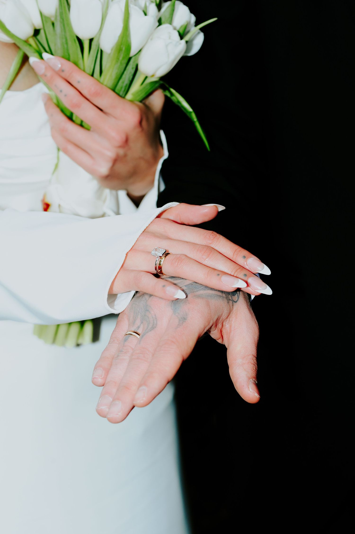 Close-up of hands holding white tulips with wedding rings visible against black background.