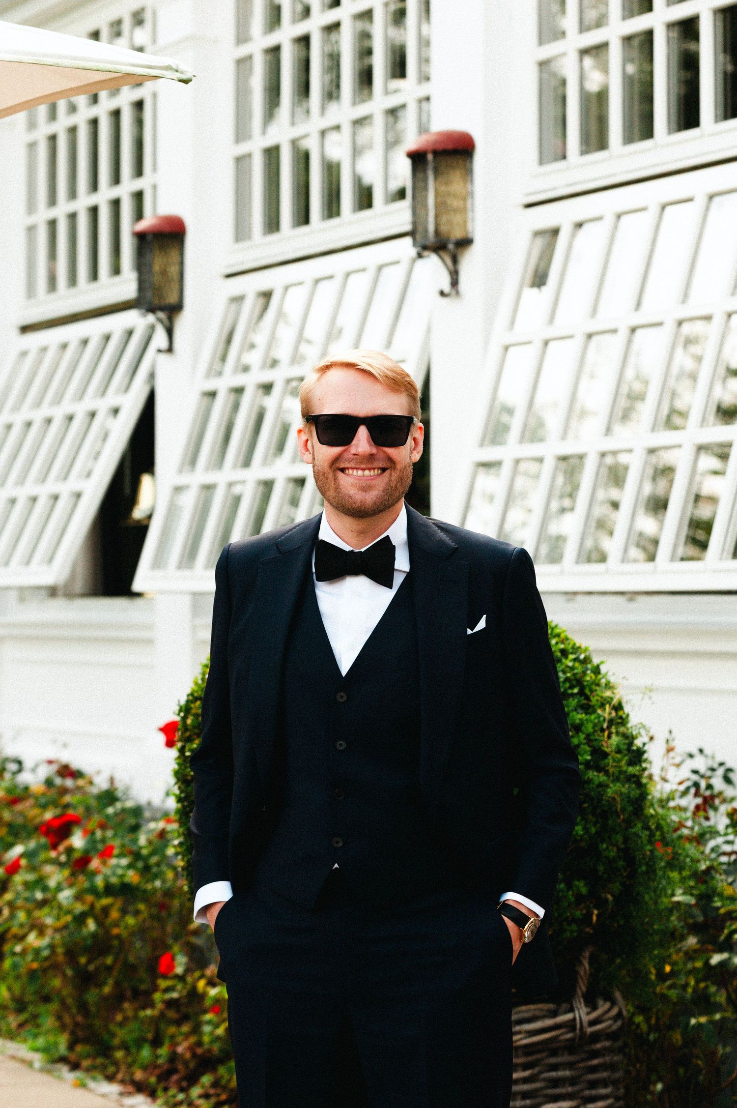 Wedding party member in black suit and sunglasses poses against white building with red flowers.