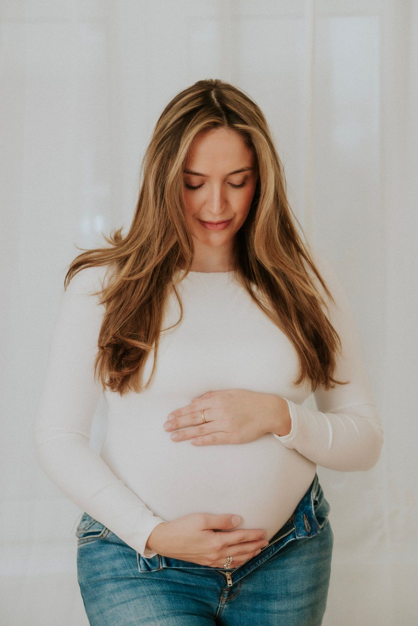 A person in a white sweater and jeans poses against a light background while cradling their belly.