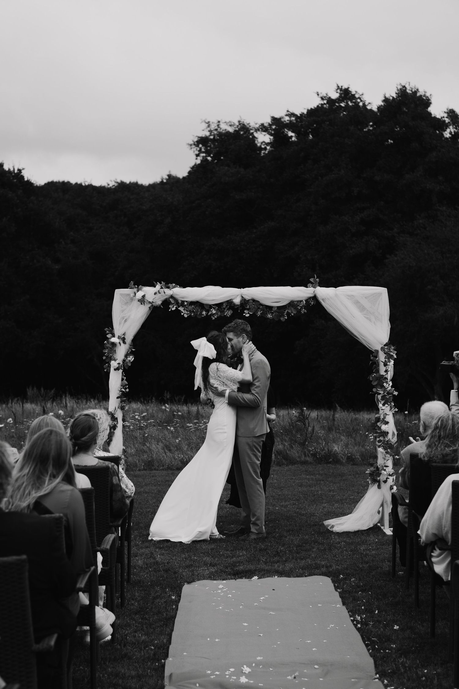 Wedding ceremony under a floral arch with guests watching in an outdoor garden setting.
