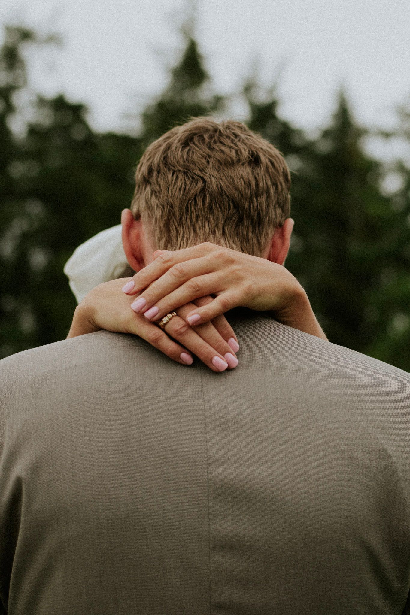 Bride's hands with wedding ring wrapped around groom's neck from behind showing intimate moment.