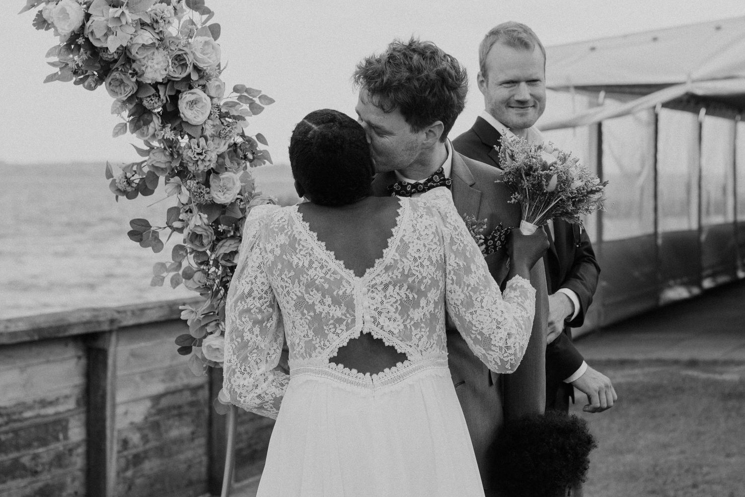 Black and white wedding ceremony photo with floral arch and water view in background.