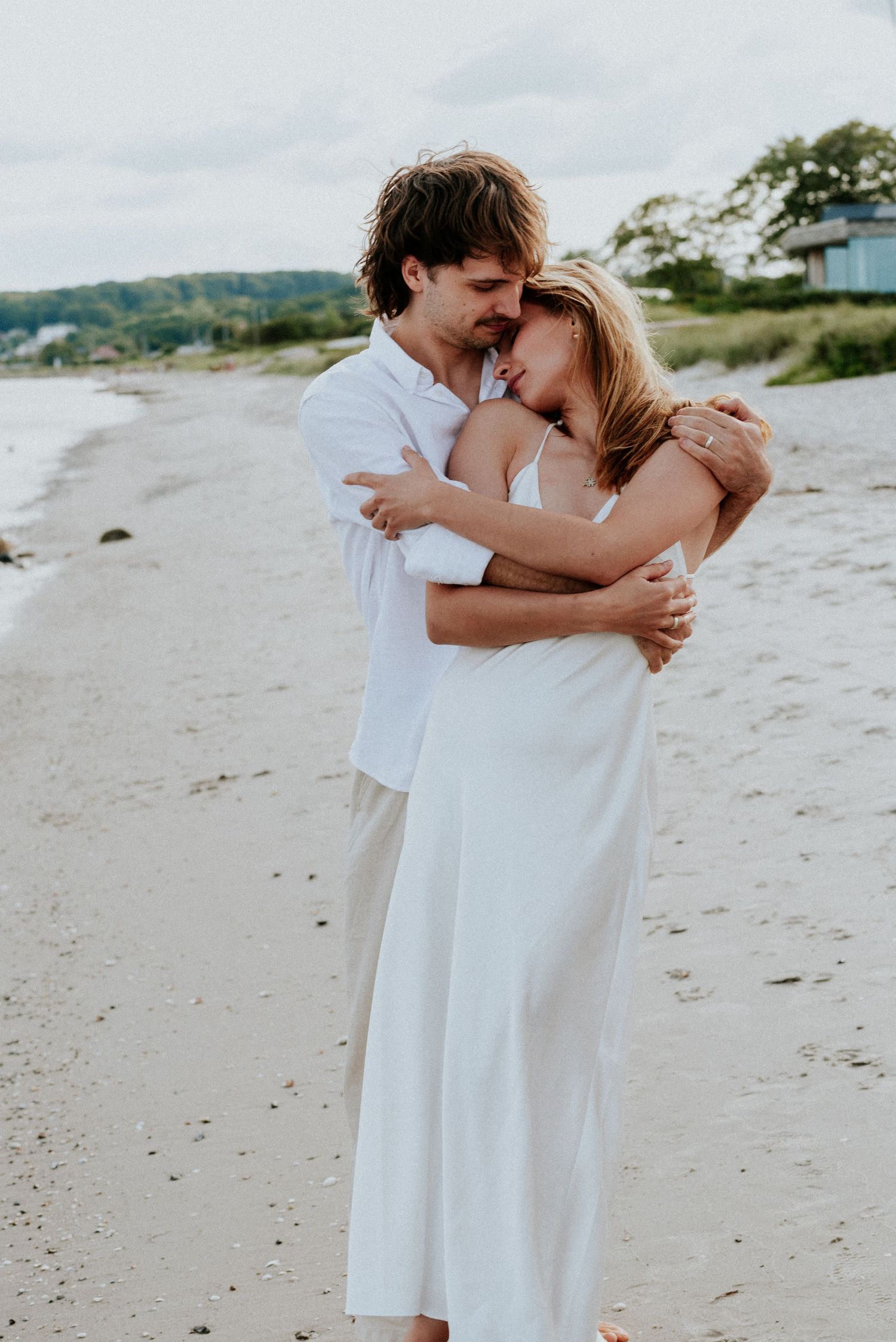 A couple in white outfits shares a romantic moment together on a sandy beach at sunset.