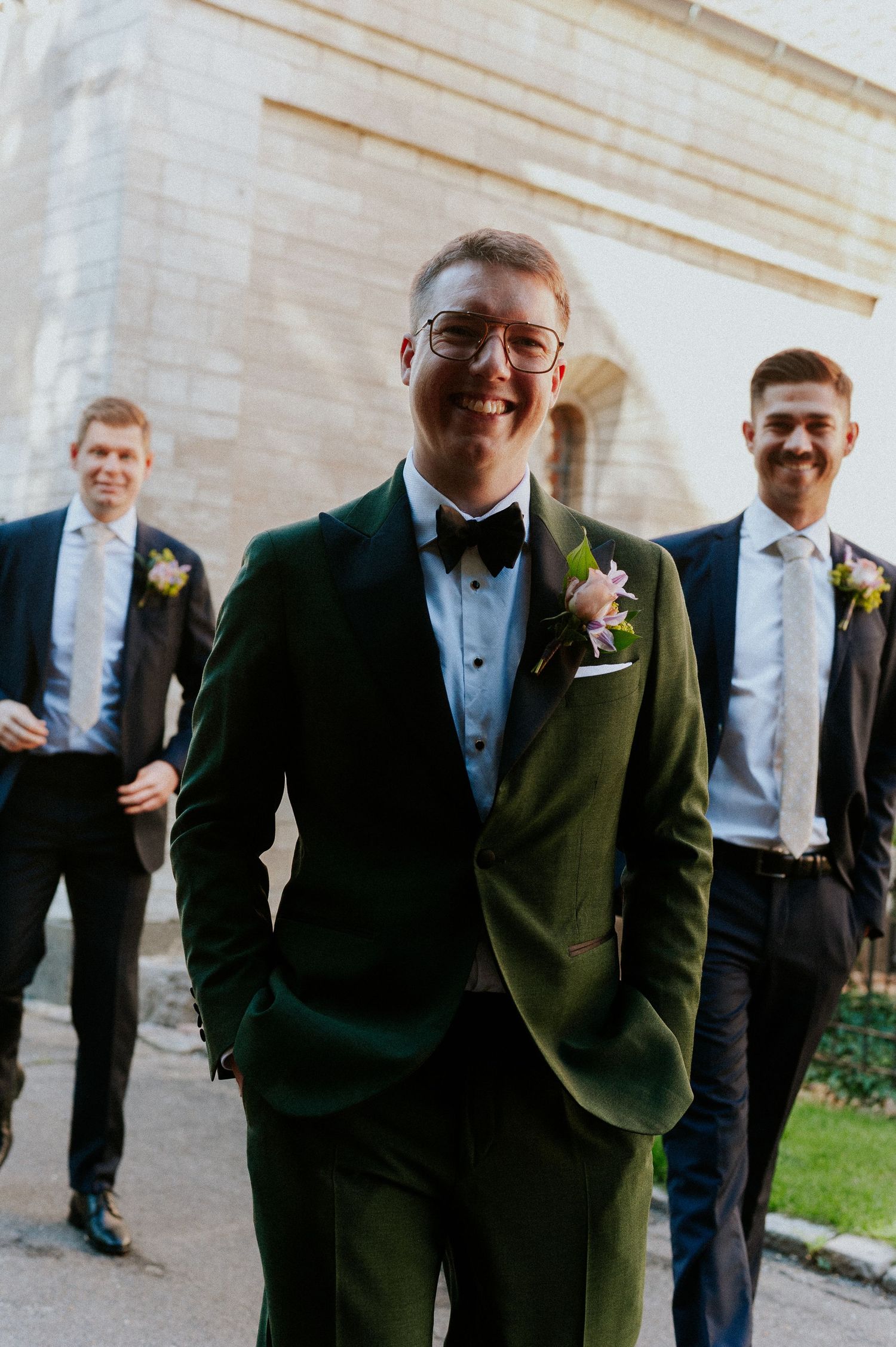 Groomsmen in dark suits and bow ties pose together outside a stone building.