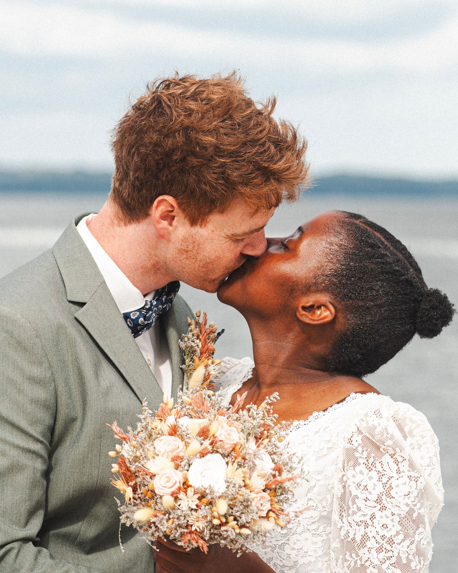 Seaside wedding portrait shows couple in gray suit and lace dress sharing romantic moment.