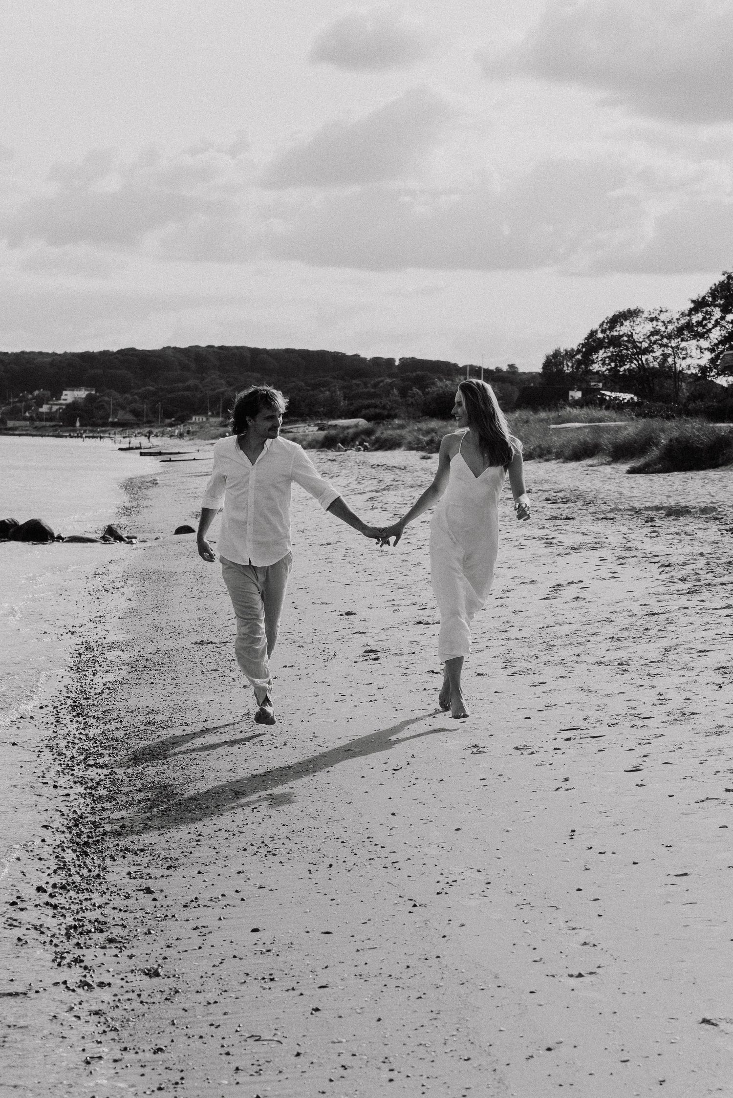 Two people in white outfits walk hand in hand along a scenic beach shoreline in black and white photography.