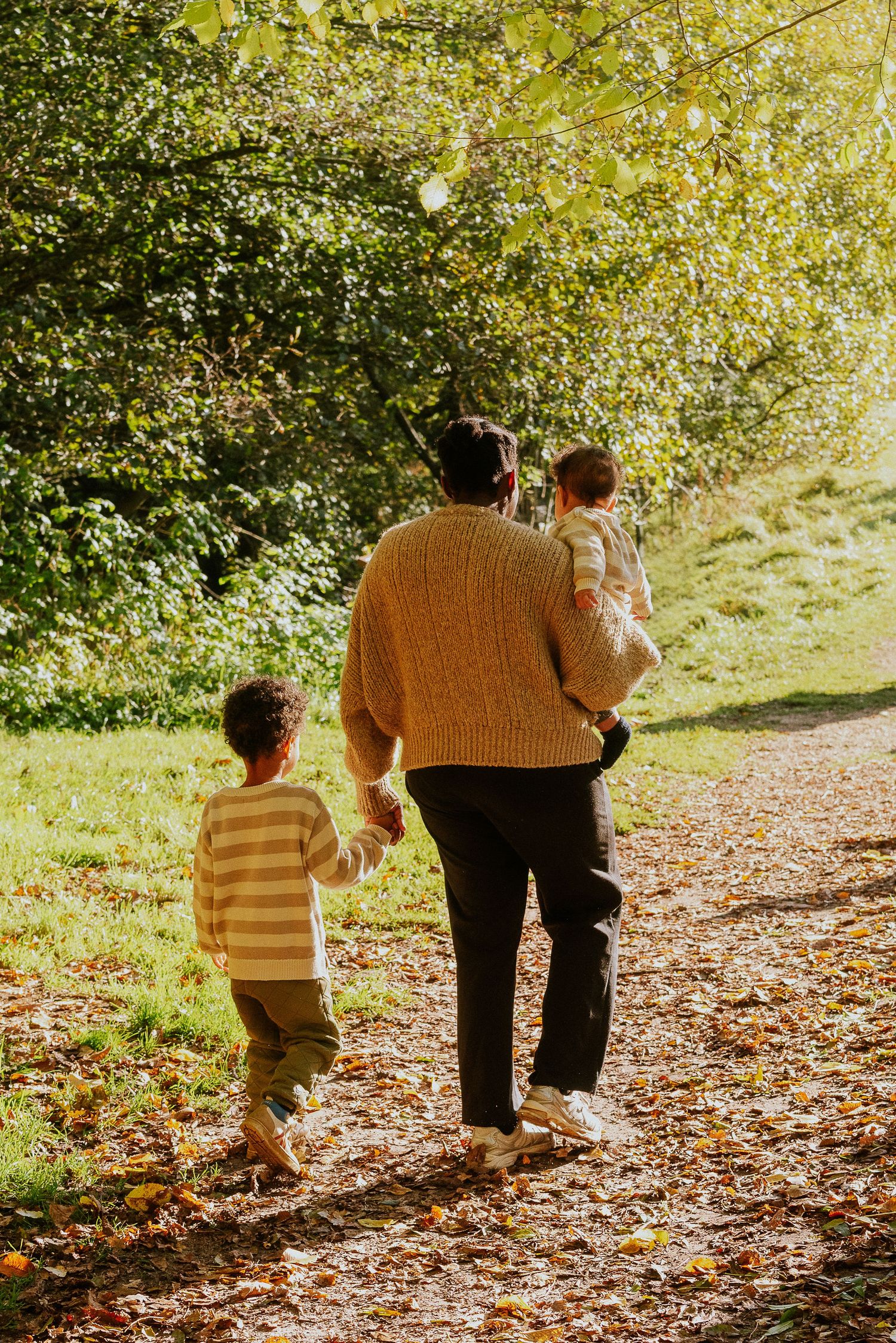 Two figures walk together on a leaf-covered autumn path through a sunlit wooded area.