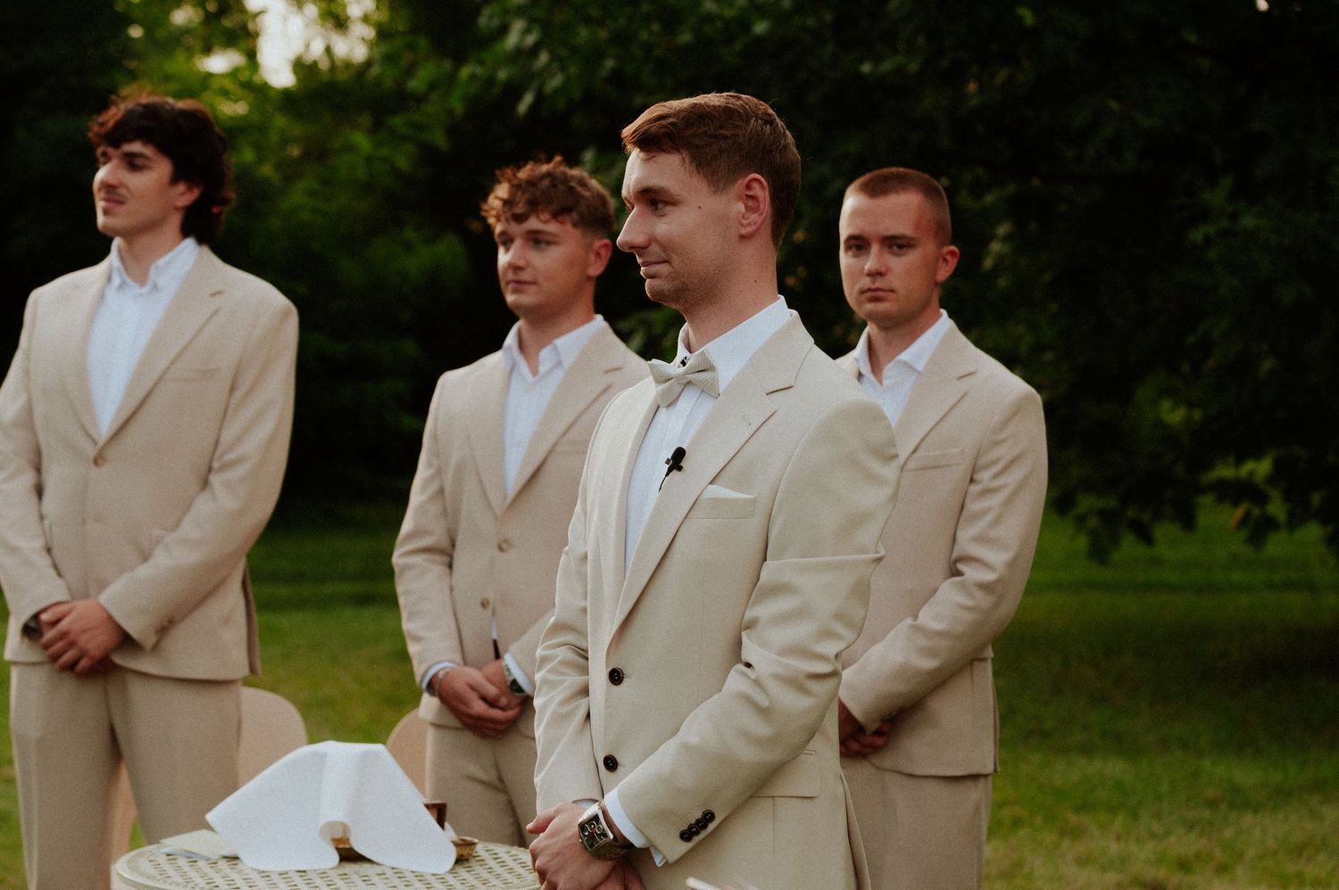 Groomsmen wearing light beige suits stand together outdoors during a wedding ceremony.