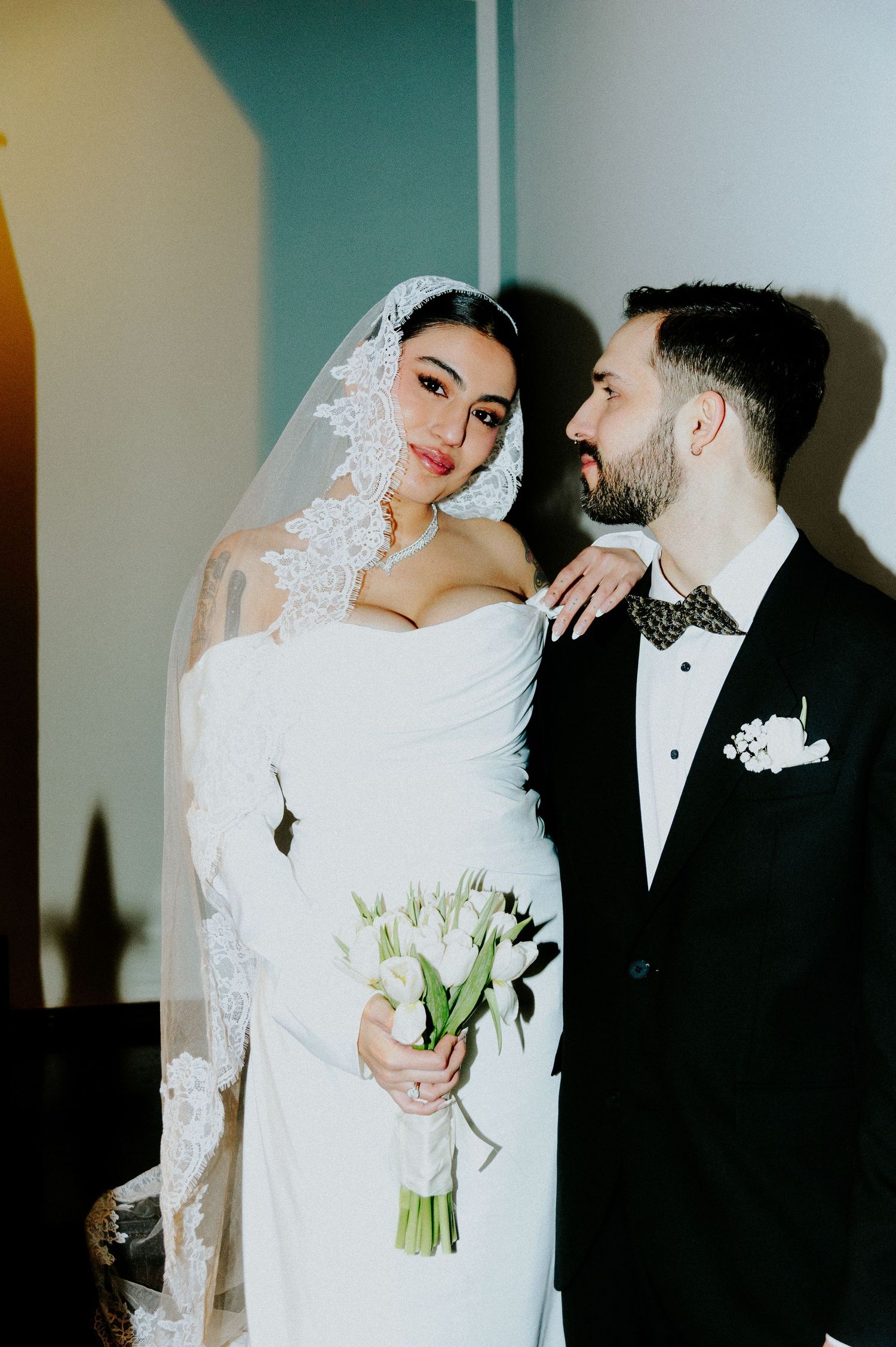 Romantic wedding portrait of couple in formal attire with white tulip bouquet against colorful wall.