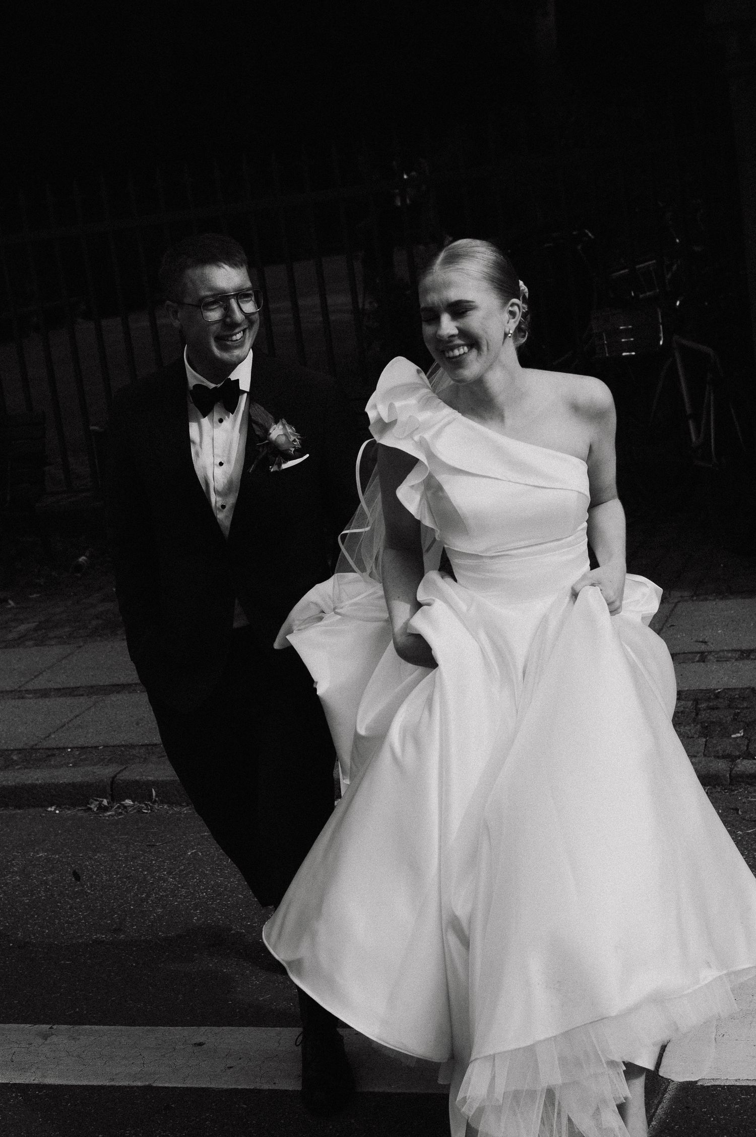 A black and white wedding photo showing a couple, with the bride in an elegant one-shoulder gown and groom in a tuxedo.