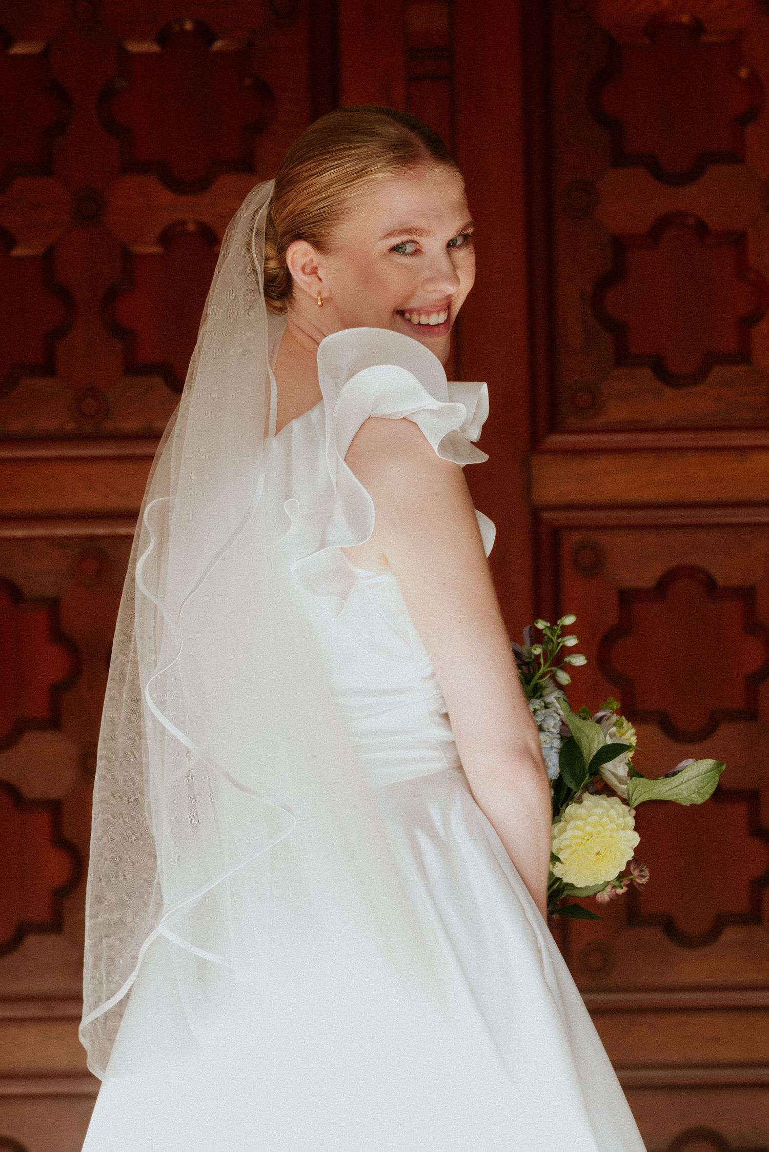 A bride in a white wedding dress and veil smiles while holding a bouquet against an ornate wooden backdrop.