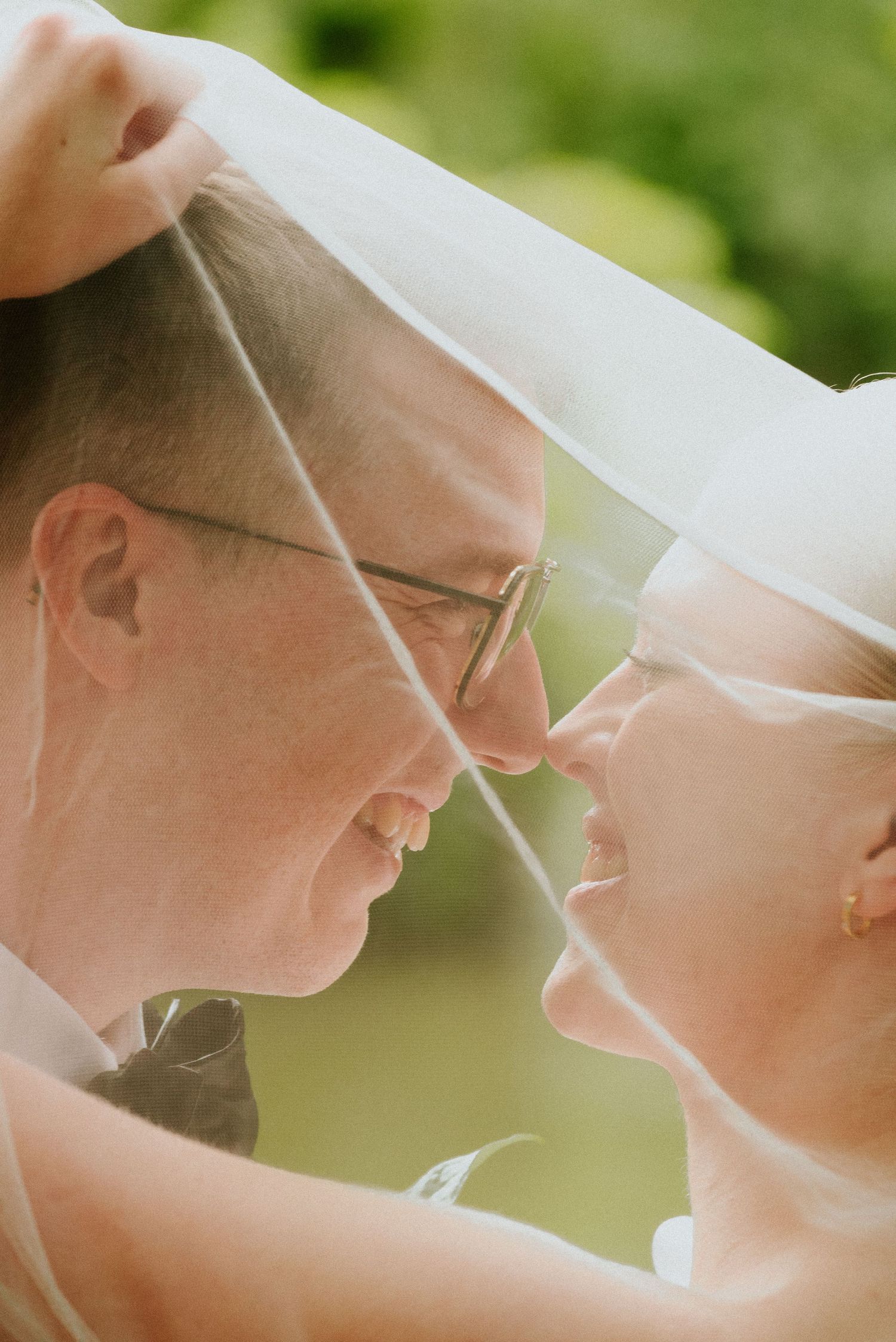 A romantic wedding couple shares an intimate moment under a white veil in soft natural lighting.