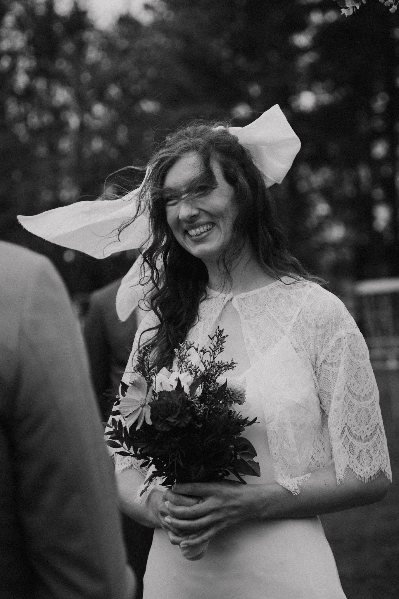 A joyful bride in a lace wedding dress holds a bouquet while laughing outdoors in black and white.