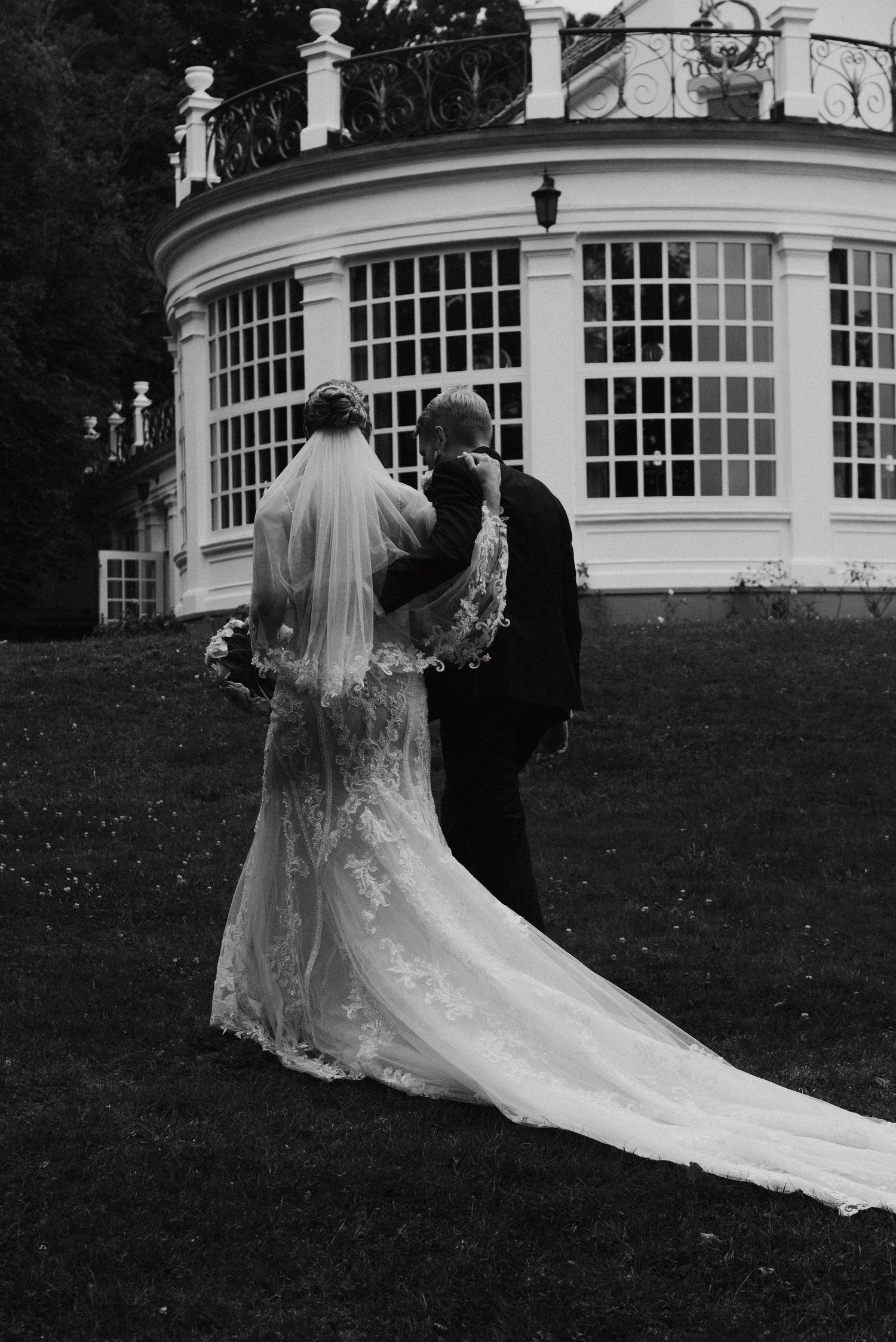 Black and white photo of couple walking near elegant curved building with trailing wedding dress.