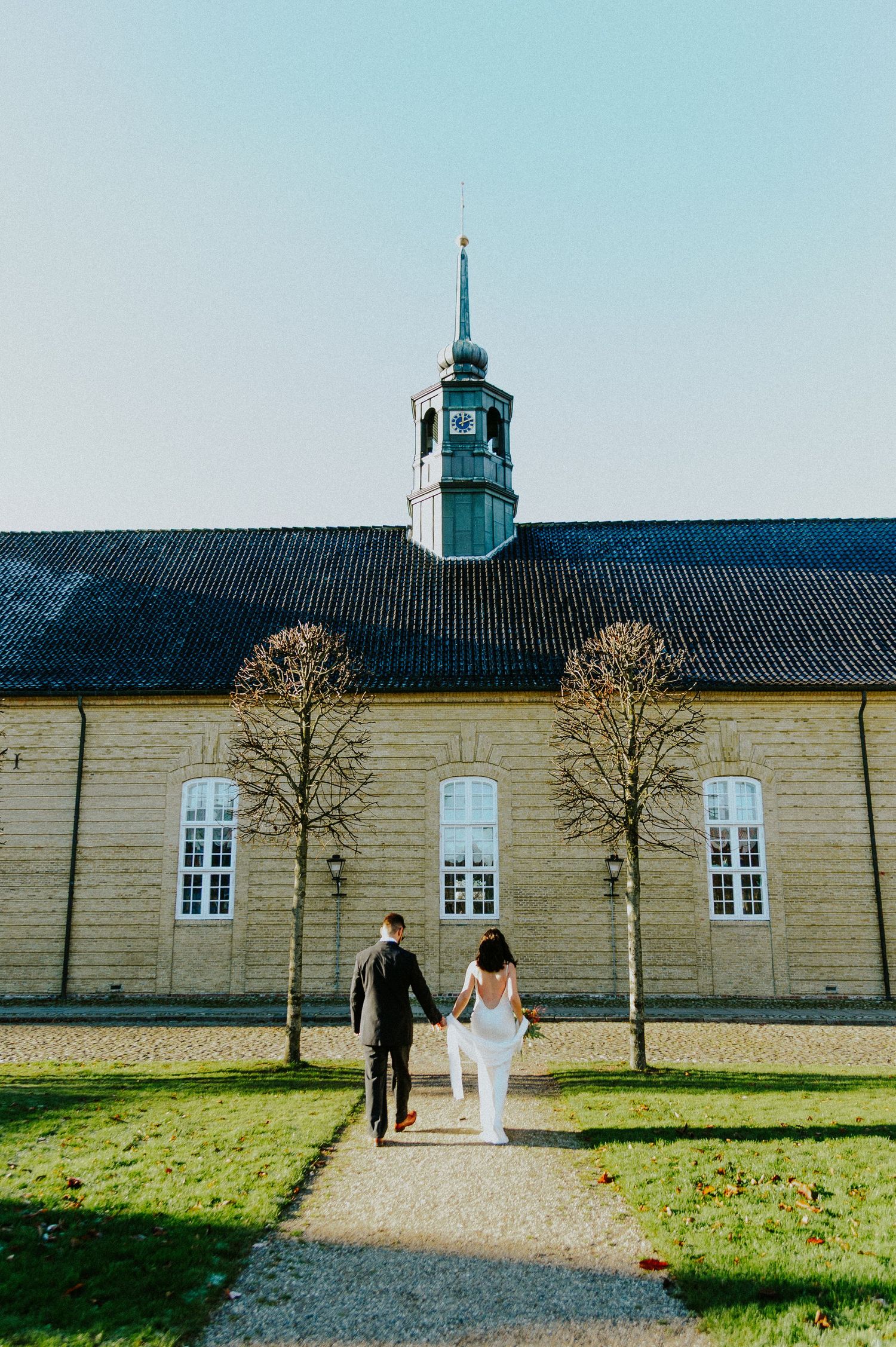 A couple walks along a gravel path toward a historic building with white walls and a blue roof.