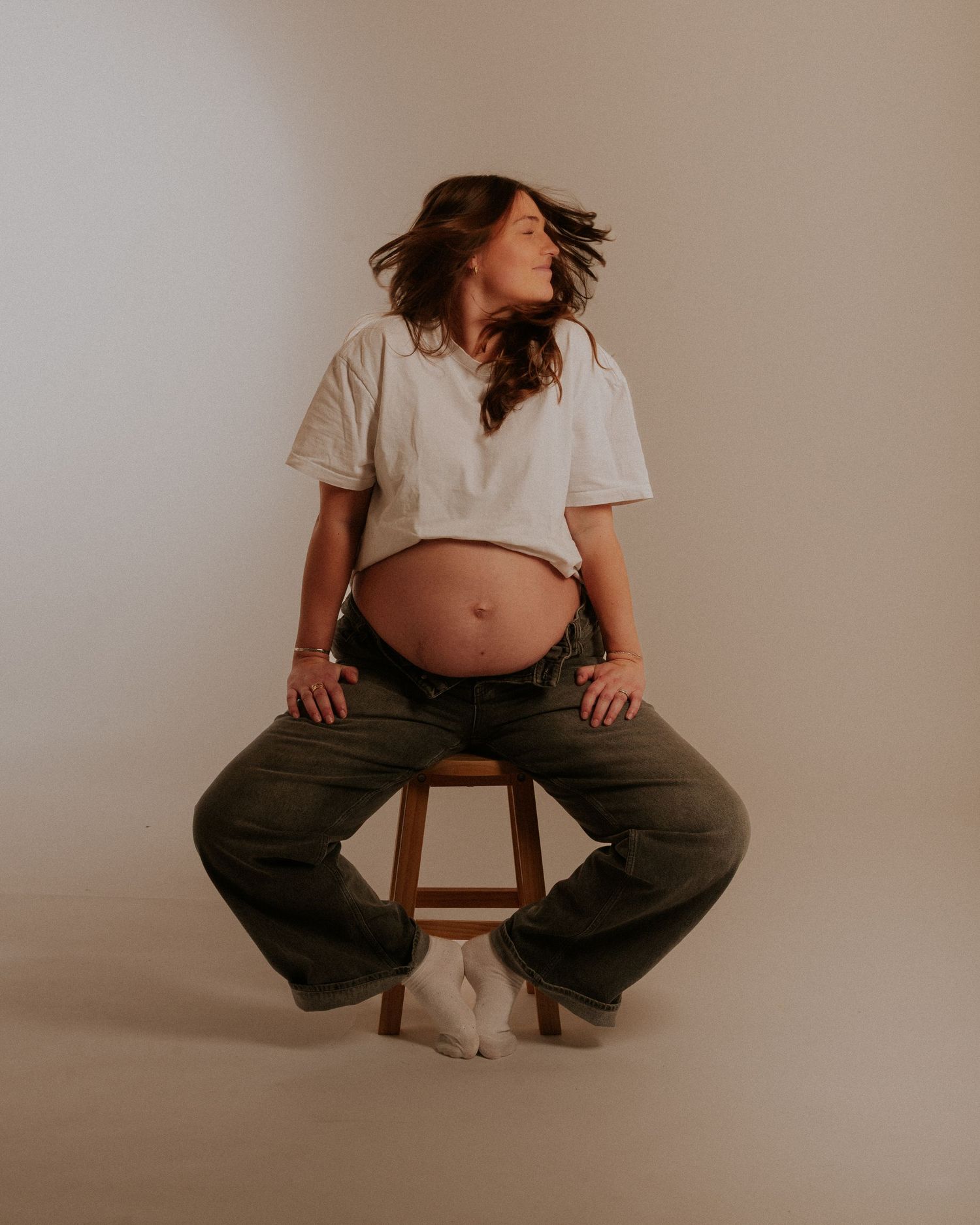 Artistic maternity photo showing an expectant mother in a crop top and cargo pants sitting on a wooden stool against neutral backdrop.