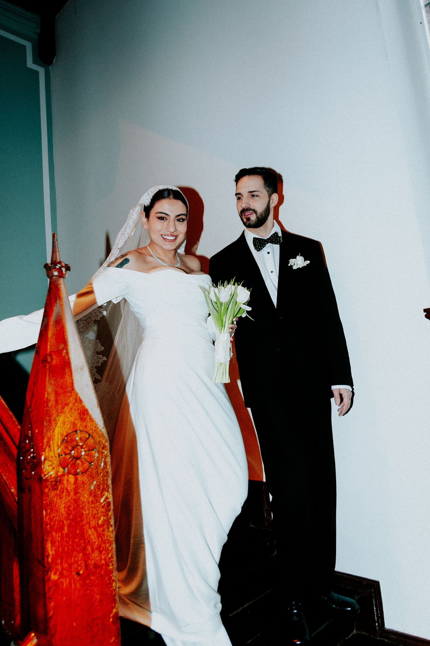 A bride and groom pose together against a white wall with a striking red art installation in the foreground.