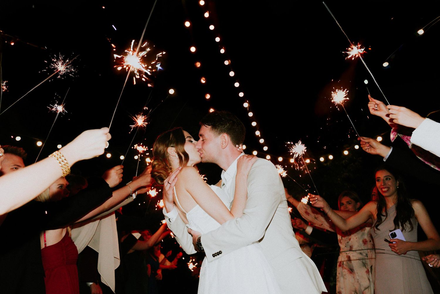 People dancing and celebrating with sparklers at a nighttime wedding reception.