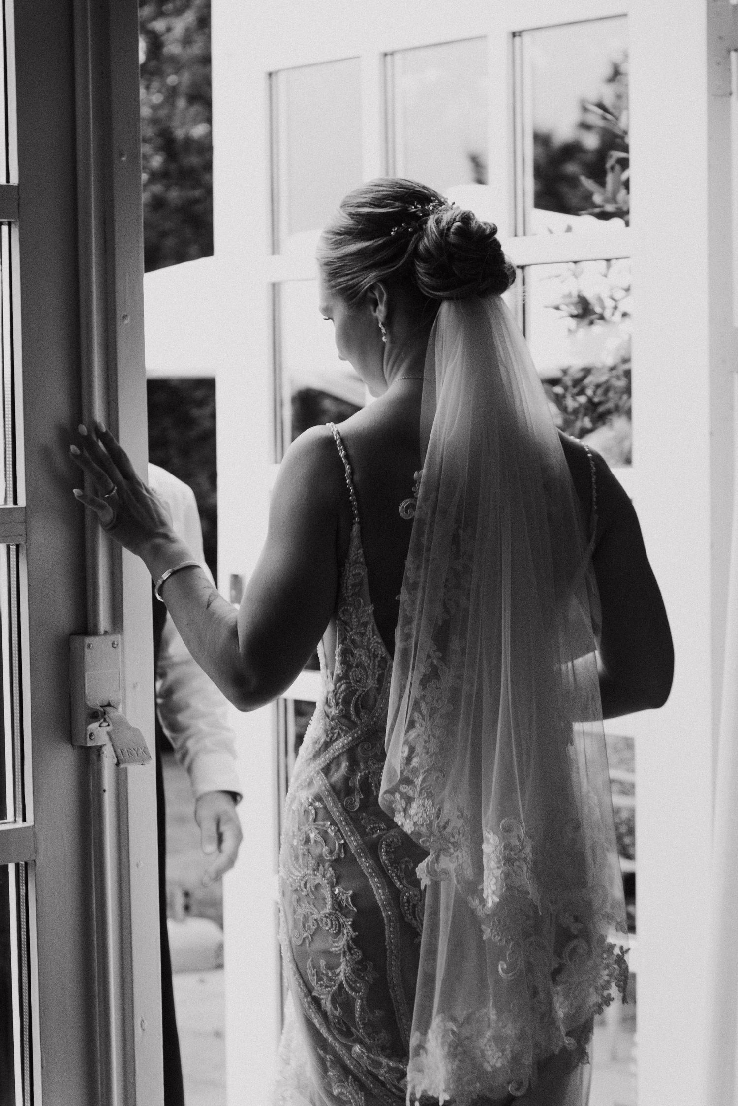 Black and white image of a bride in a lace dress looking out windows in an elegant interior setting.