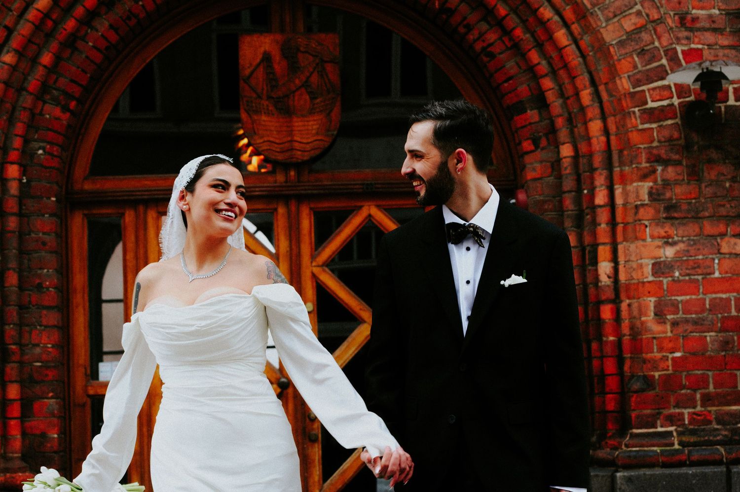 Joyful wedding moment captured under ornate red brick archway with couple in classic formal attire.