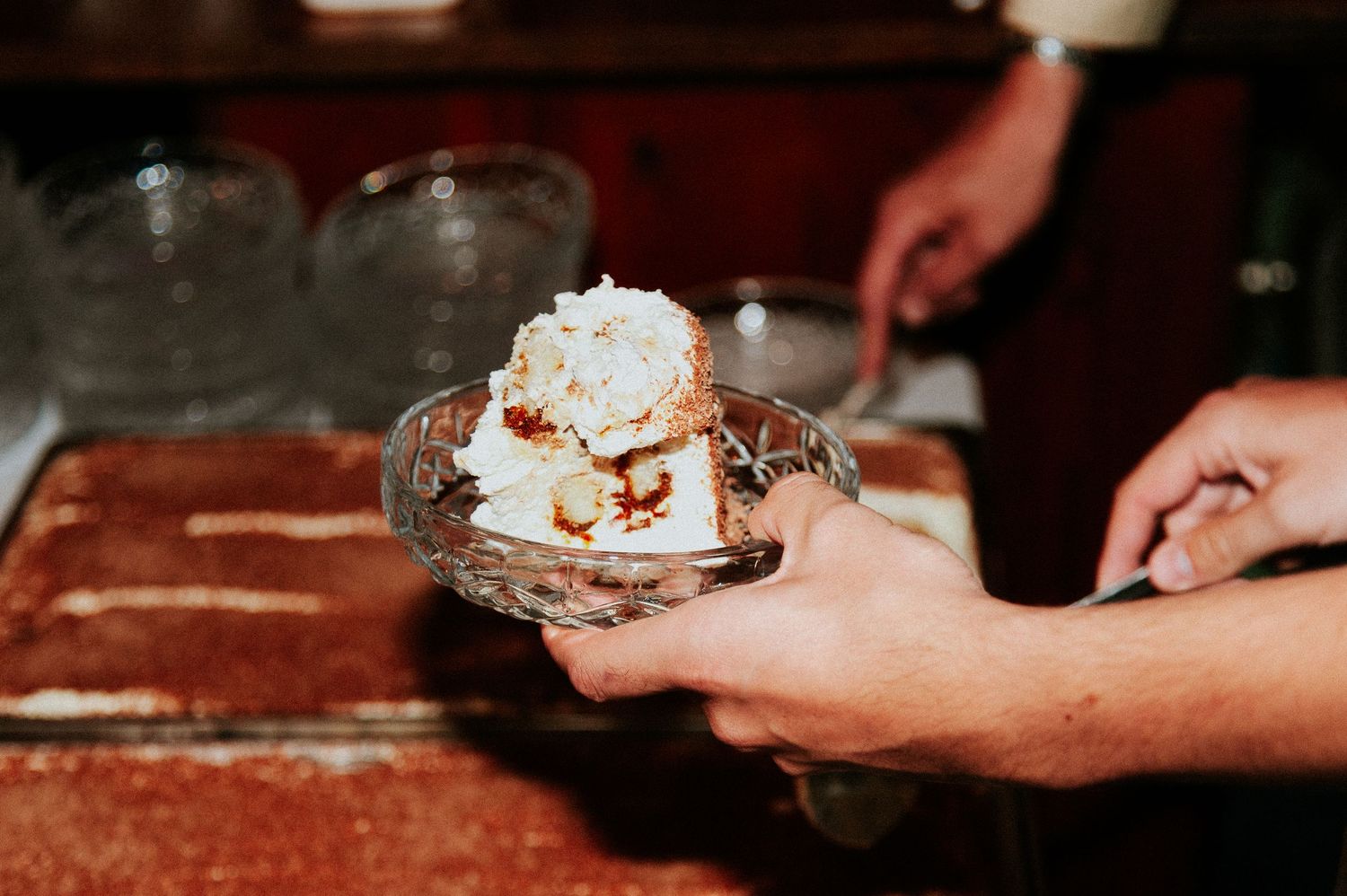 Hands serving slices of homemade pie topped with whipped cream and crumbs on rustic wooden surface.
