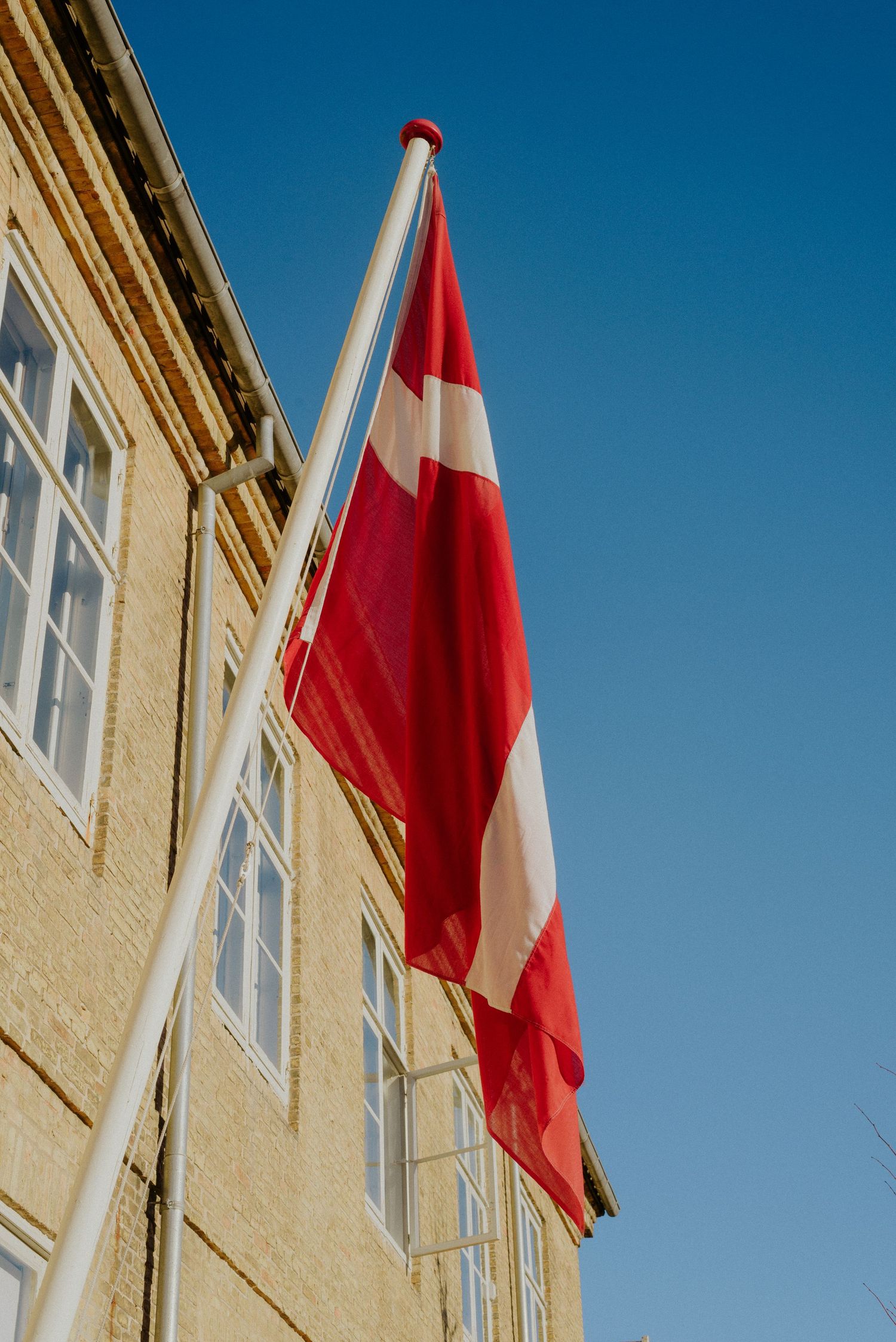 Red and white flags waving against a bright blue sky beside a yellow building facade.