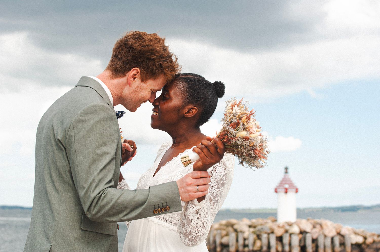 A joyful wedding couple embraces by the waterfront with a lighthouse in the background.