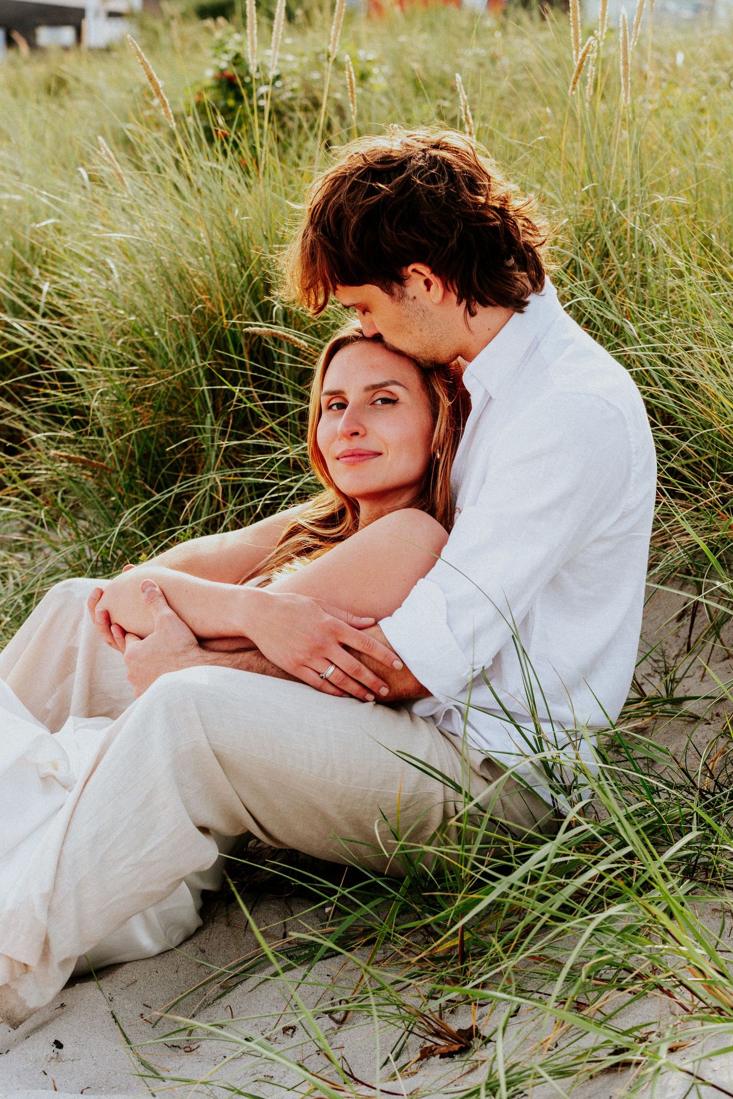 A romantic couple embraces while lying in tall beach grass on a sunny day.