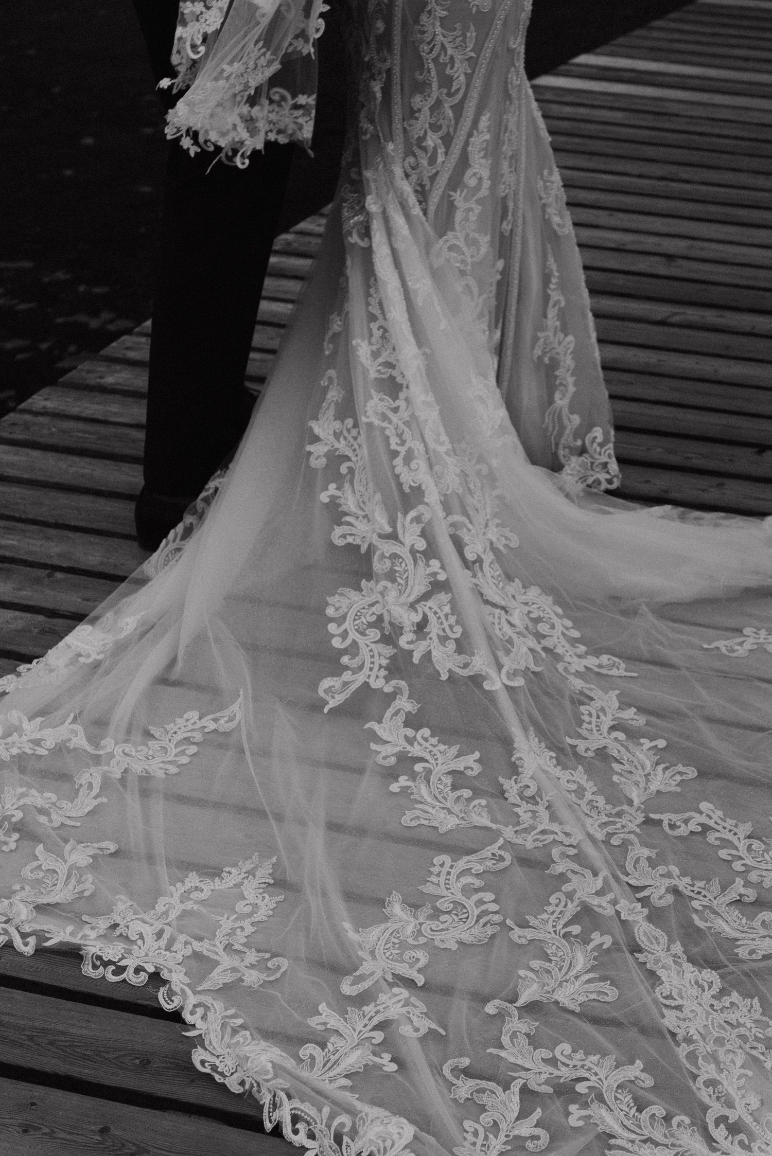 Detailed black and white shot of an ornate lace wedding dress train on wooden decking.