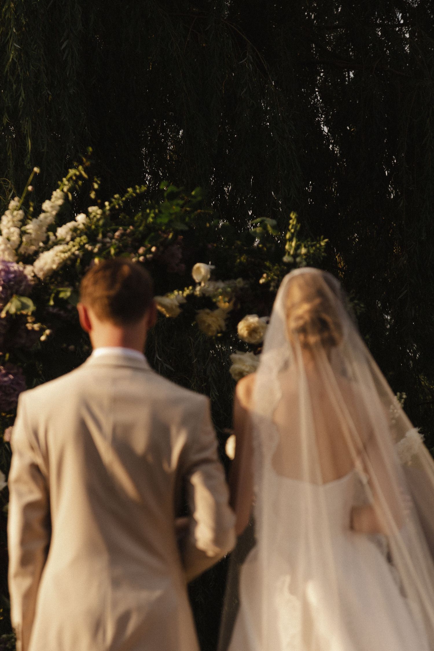 A couple in wedding attire stands before a floral arch during their outdoor ceremony.
