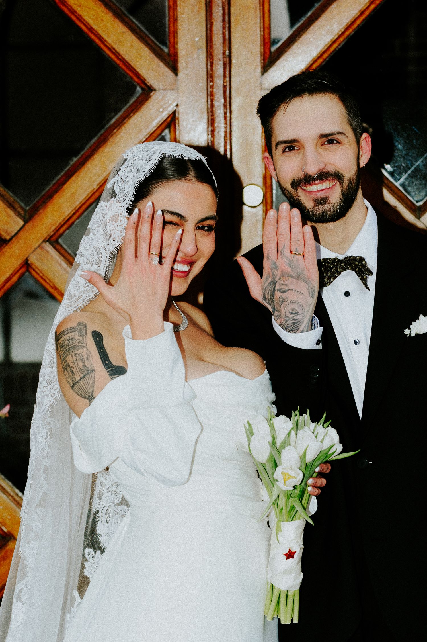 Newlyweds showing off their rings while holding white tulips against wooden door backdrop.