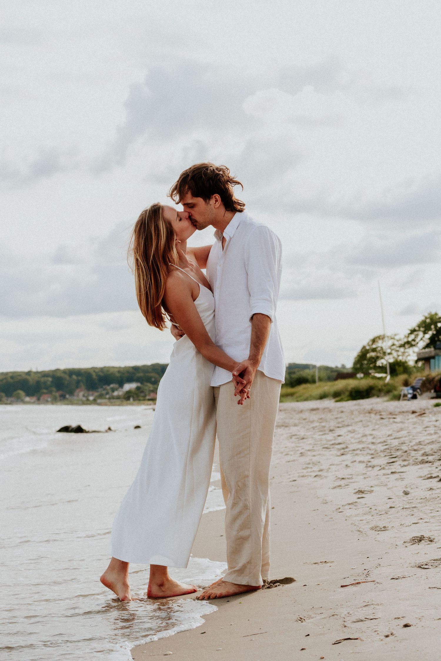 Romantic couple in white outfits embrace on a serene beach shoreline with cloudy skies in the background.
