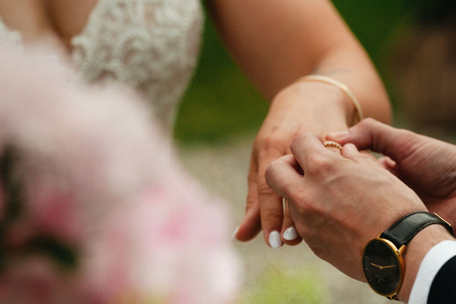 Close-up of hands exchanging wedding rings during an outdoor ceremony with lace dress and greenery in background.