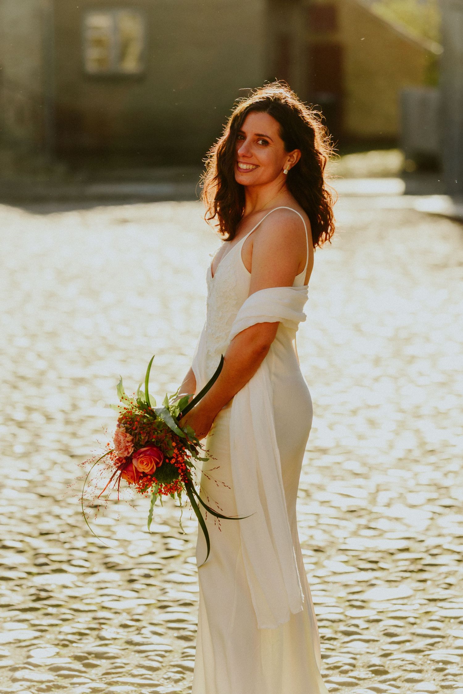 Bride in elegant white dress poses by water at golden hour holding vibrant red bouquet.