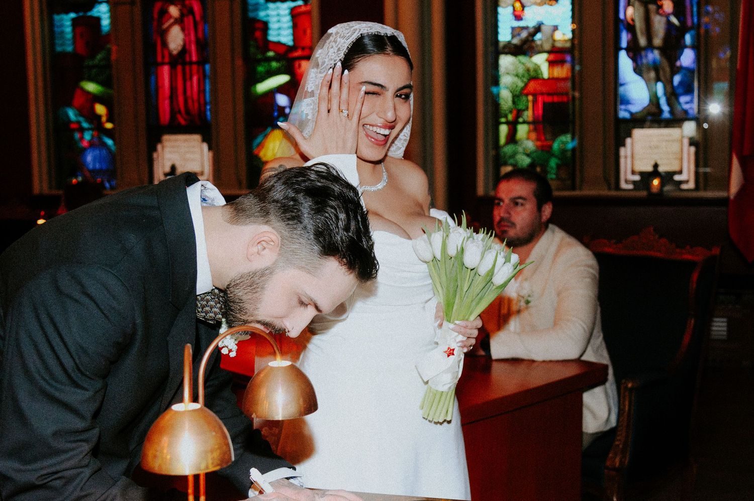 A couple signs documents at a table with a warm lamp illuminating the scene during an indoor ceremony.
