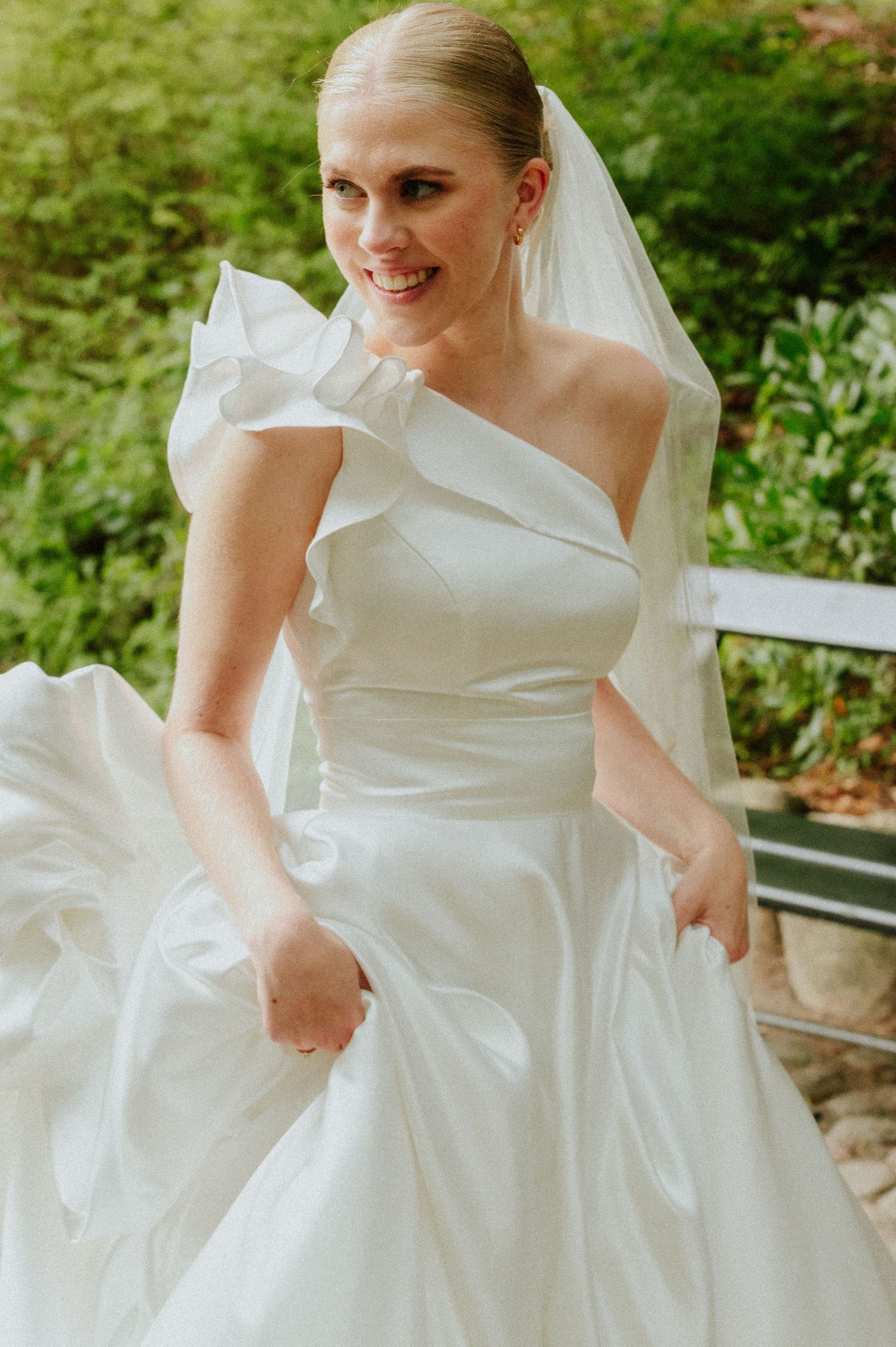 Elegant wedding dress with asymmetrical ruffled neckline and flowing skirt photographed in a garden setting.