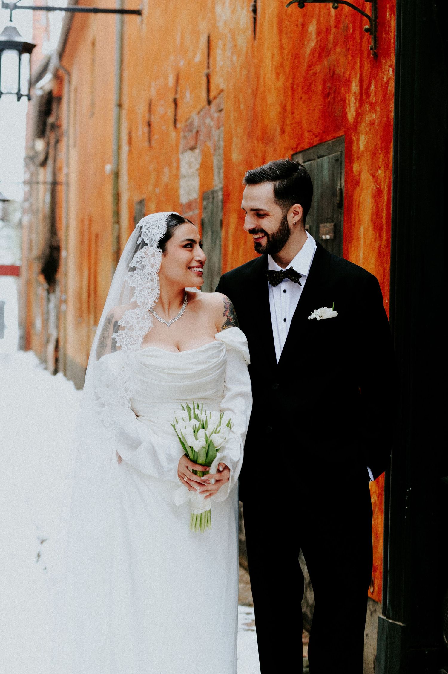 Wedding couple poses against orange wall with bride holding white floral bouquet.