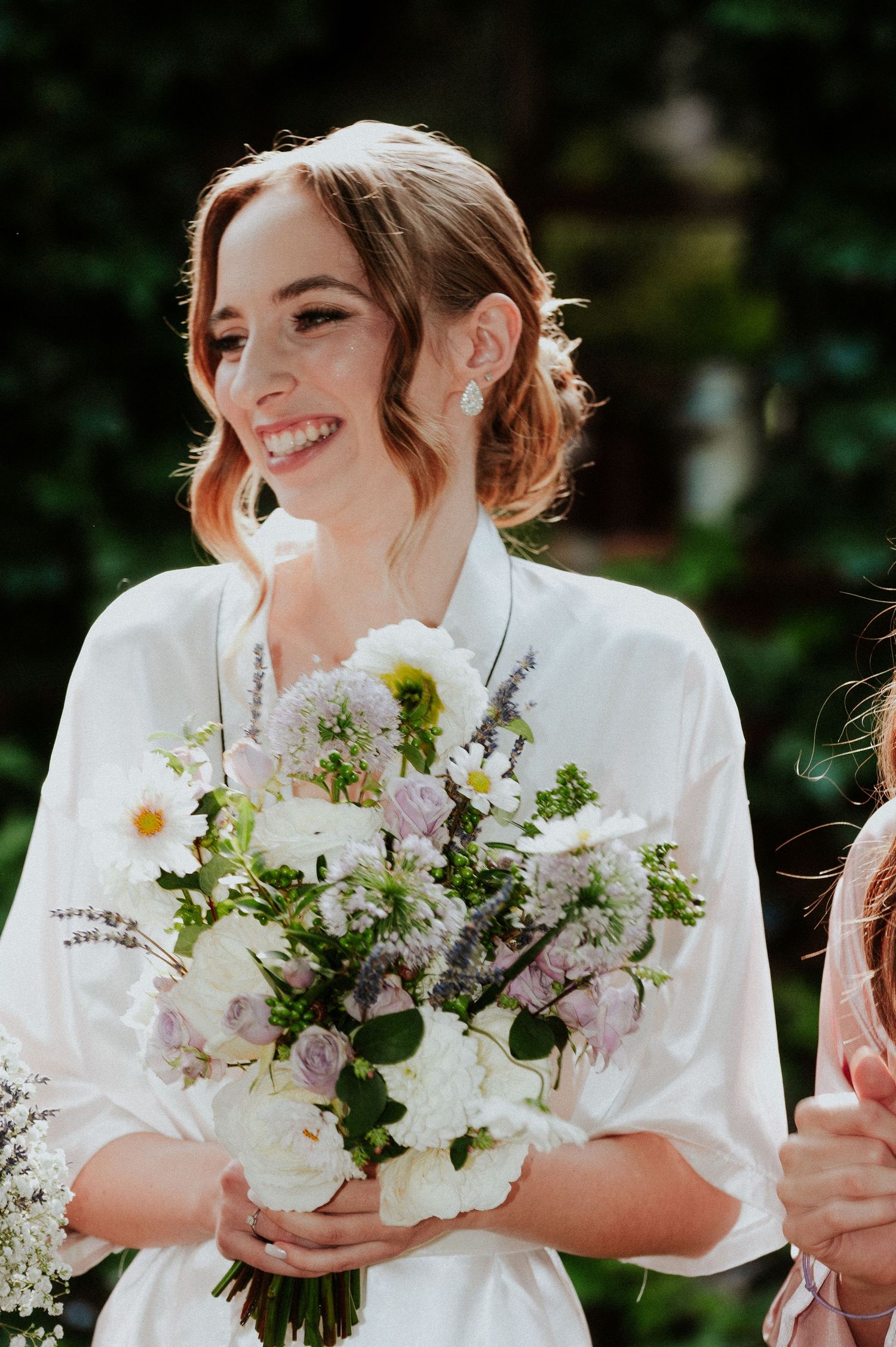 A bride in white holds a delicate bouquet of white flowers and greenery while smiling outdoors.