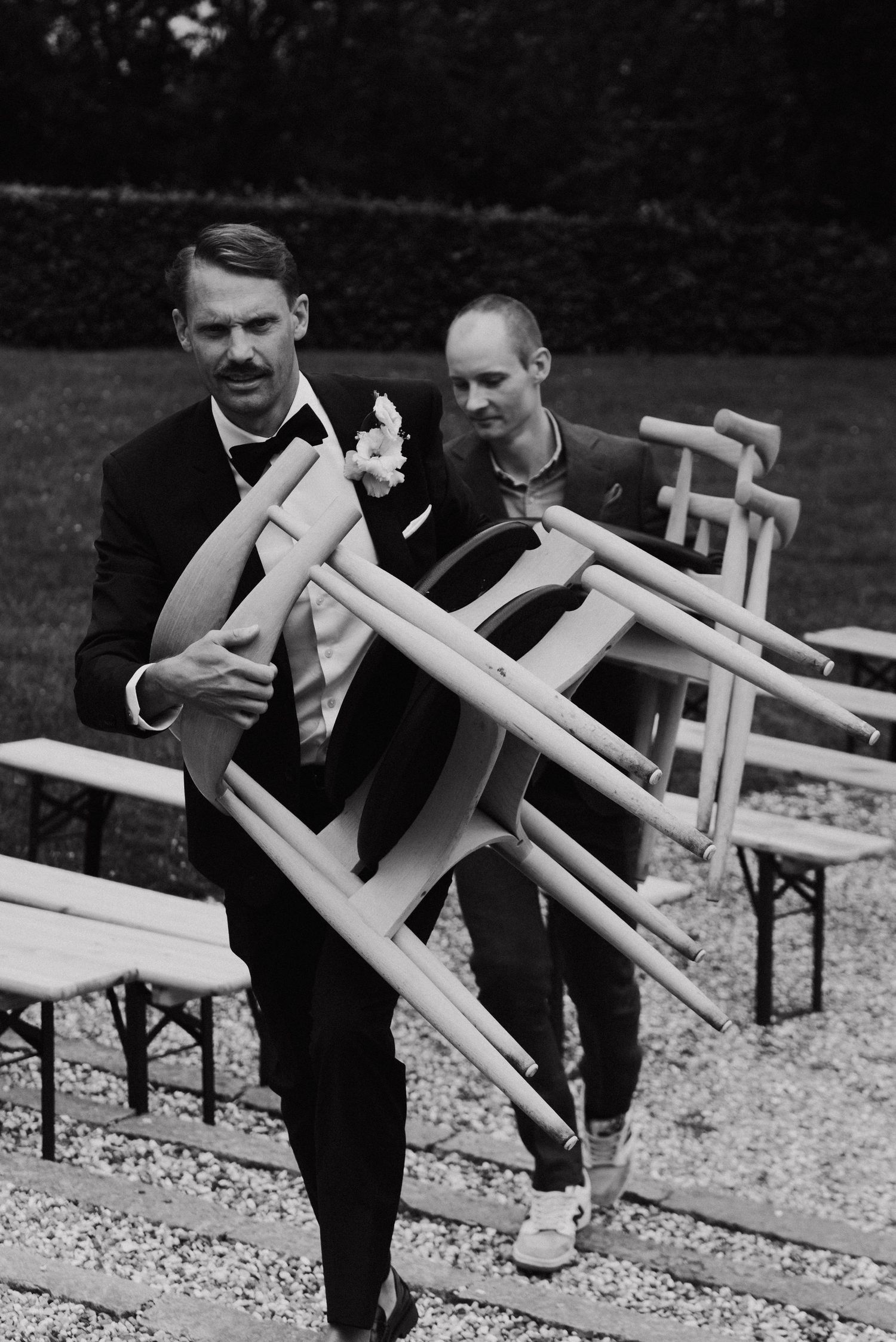Men in suits carrying white wooden chairs across a gravel surface in a black and white photograph.