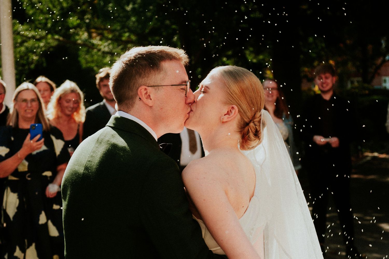 A romantic wedding kiss outdoors with guests in the background.