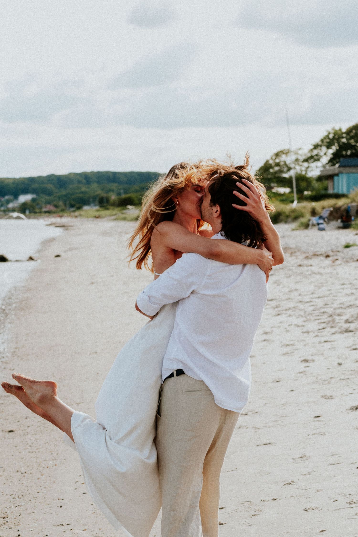 A couple in white and beige outfits embrace playfully on a sandy beach with coastal scenery in the background.