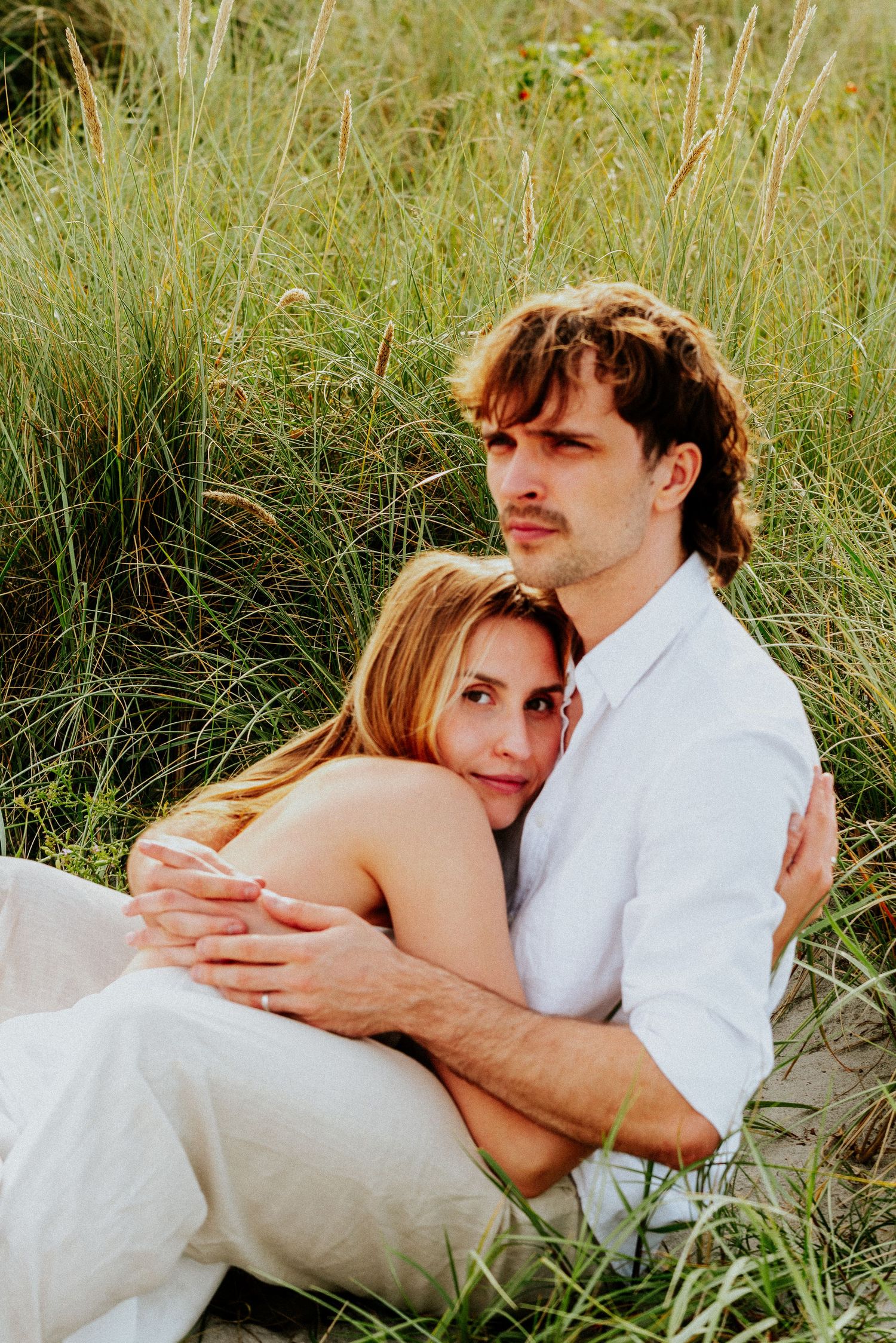 A couple embraces tenderly while sitting in tall grass during a romantic outdoor photoshoot at sunset.
