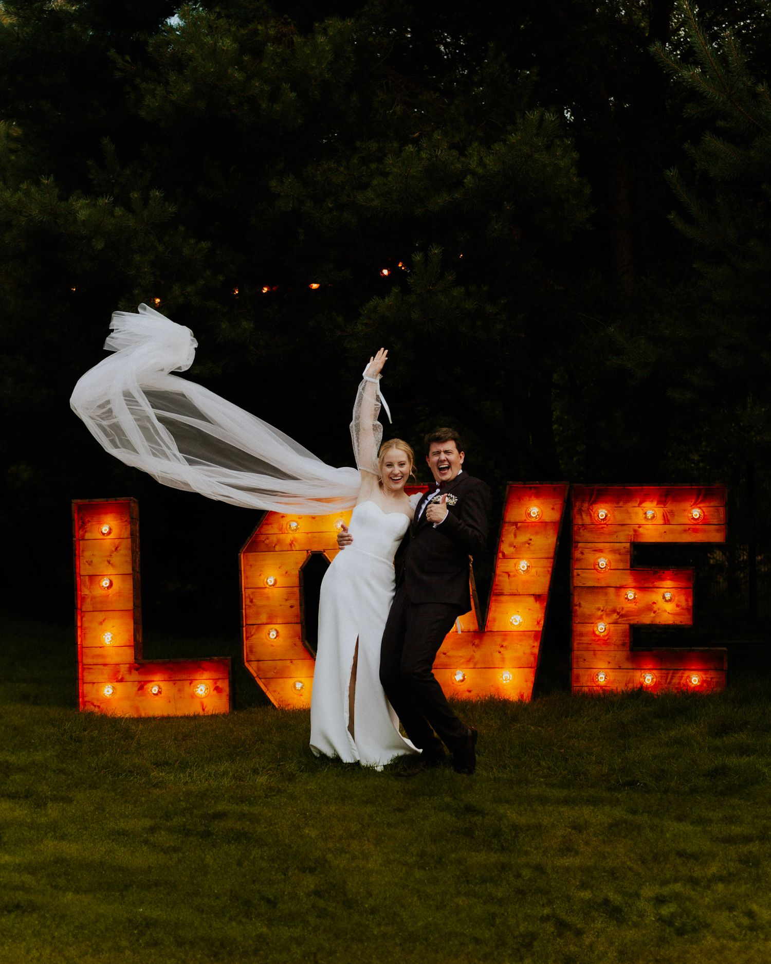 Couple poses playfully in front of illuminated LOVE marquee letters at night.