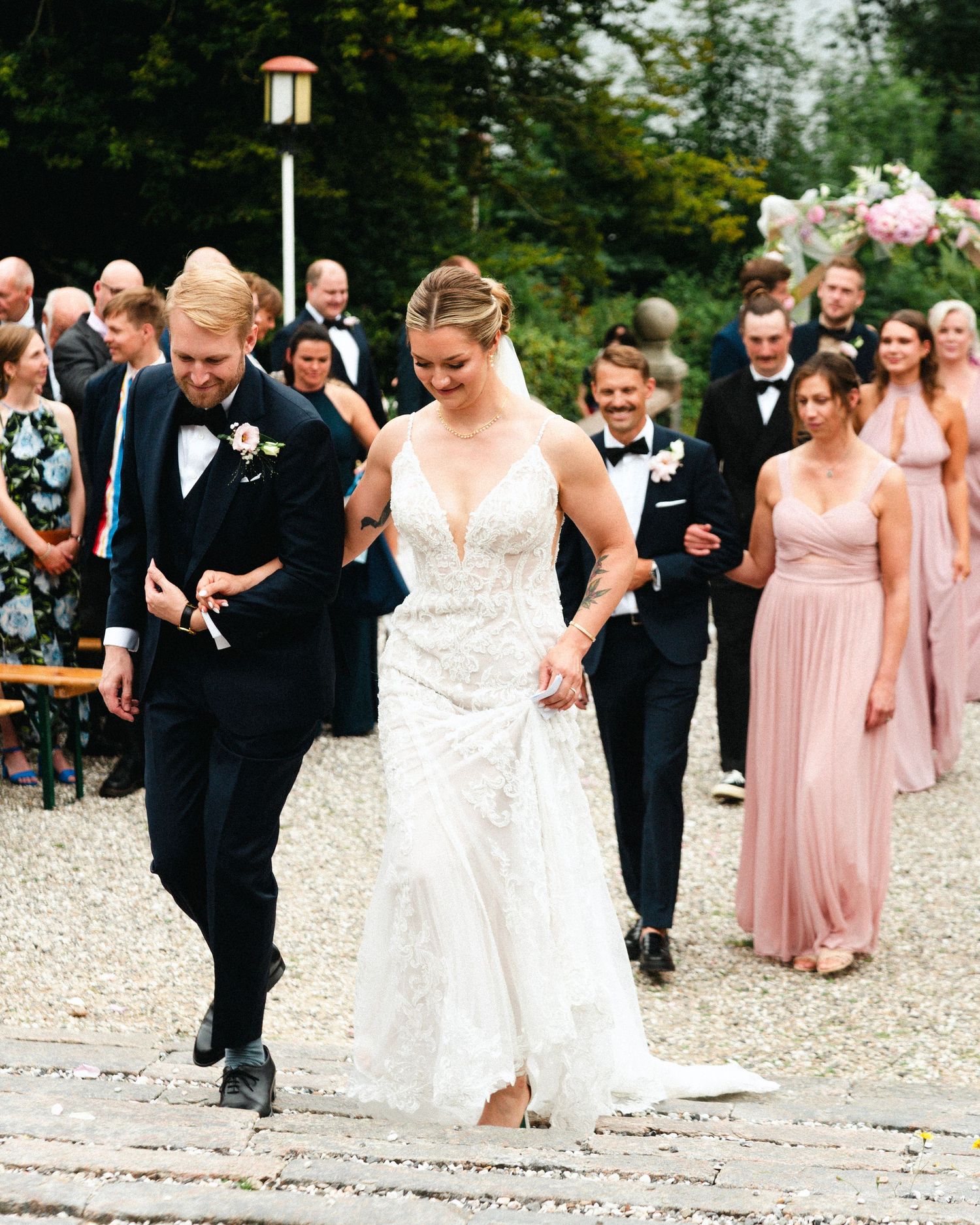 Wedding party walks down a gravel path with bridesmaids in pink dresses and groomsmen in black suits.