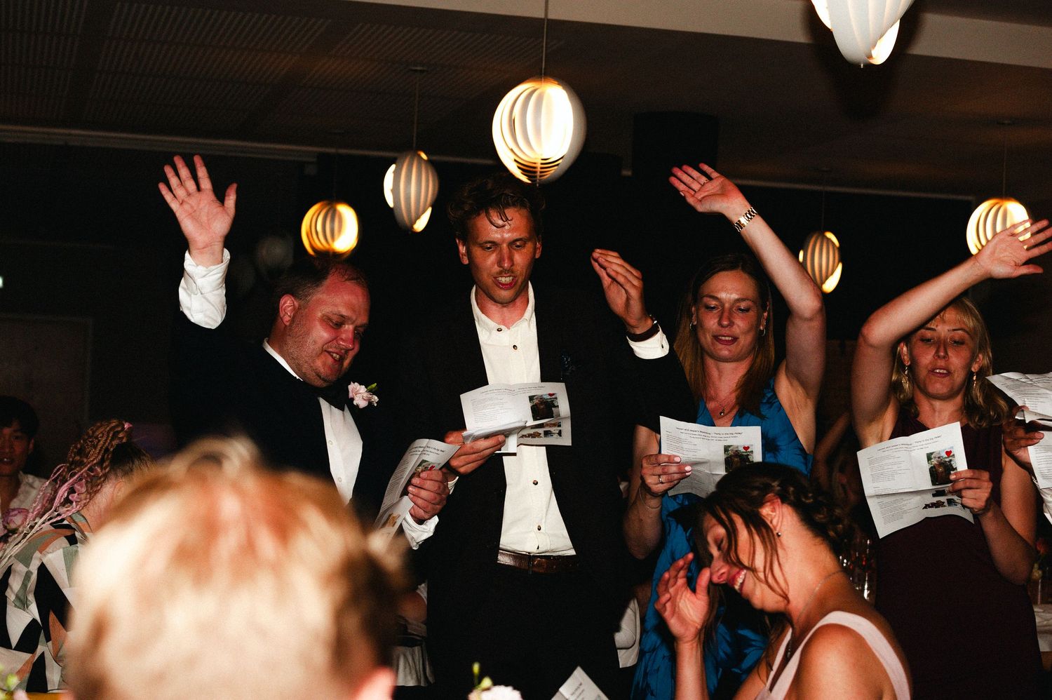 Wedding guests dance enthusiastically with raised arms at a dimly lit reception party.