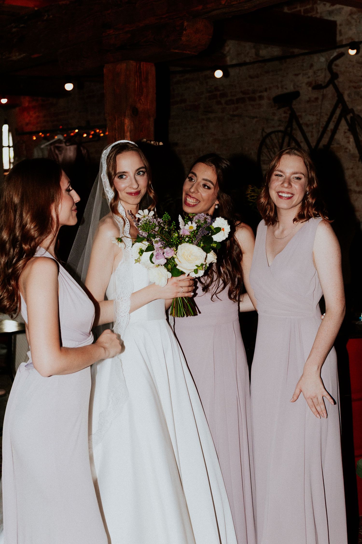 Wedding party in white and blush pink dresses gather together in a rustic indoor venue.